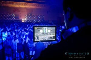 DJ using a laptop to mix music at a crowded, dimly lit dance event.