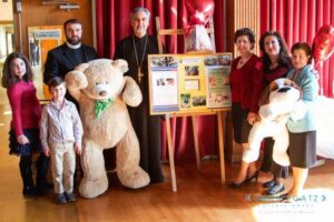 A group of seven people stands indoors with a large teddy bear and a large plush dog. There is a display board in the background with photos and text.