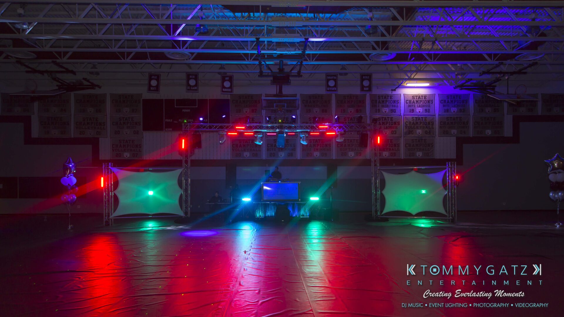 Broadneck-HC-FINAL-2017-BLOG-LoRes-WM-1 - TOMMY GATZ ENTERTAINMENT Empty gymnasium decorated for an event with colorful stage lights and two projector screens, branding for "Tommy Gatz Entertainment" visible at the bottom right.