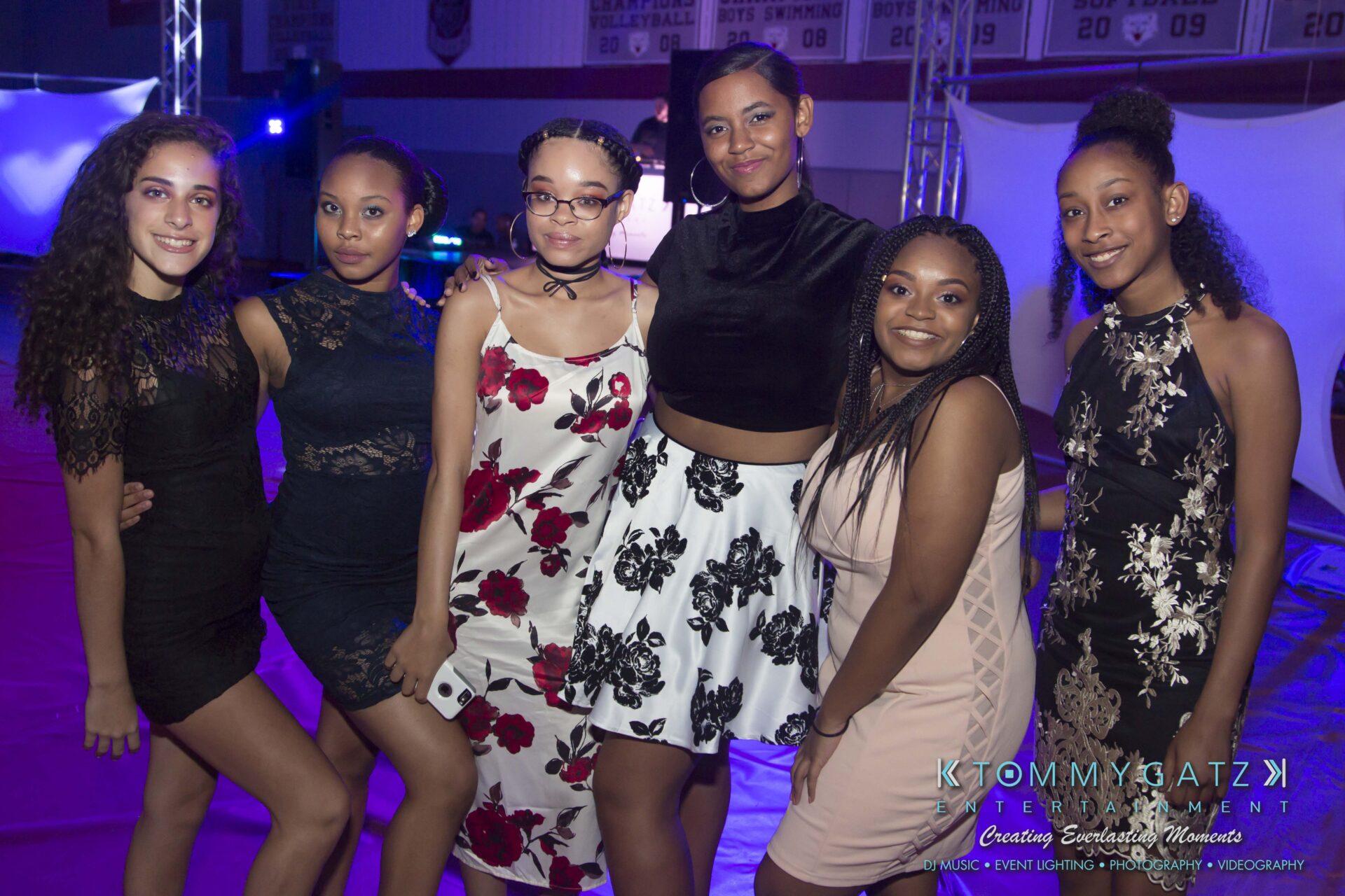 Broadneck-HC-FINAL-2017-BLOG-LoRes-WM-23 - TOMMY GATZ ENTERTAINMENT Six women pose together at an indoor event, wearing semi-formal dresses with floral and solid patterns. Blue lighting sets a festive atmosphere.
