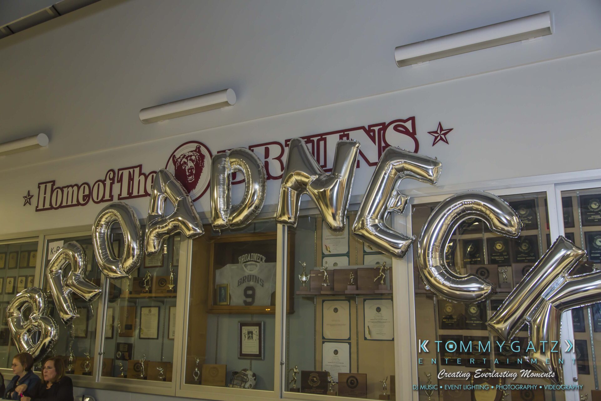 Broadneck-HC-FINAL-2017-BLOG-LoRes-WM-3 - TOMMY GATZ ENTERTAINMENT Silver balloons spelling "BROADNECK" in front of a trophy display and a sign reading "Home of The Bruins.