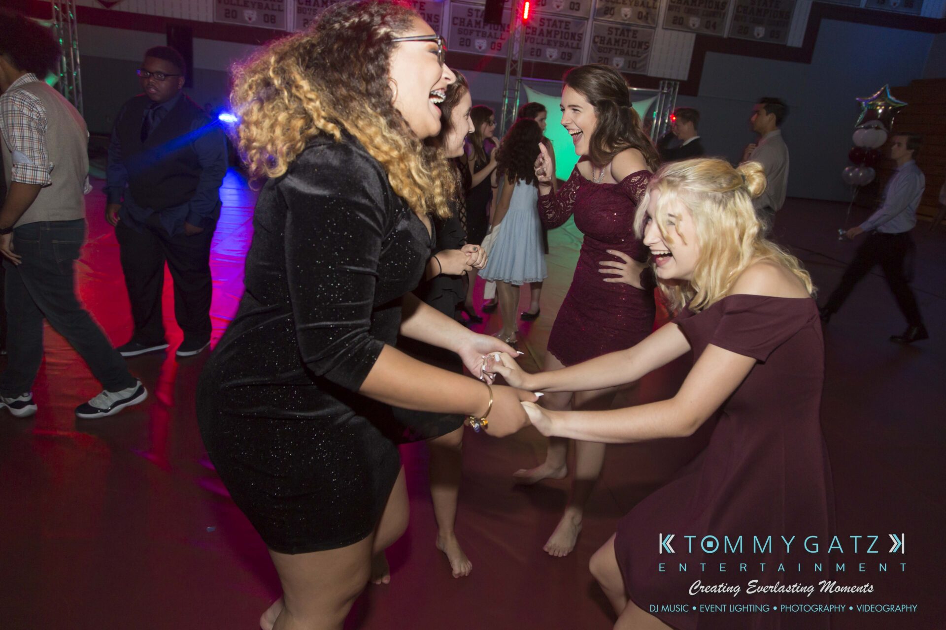 Broadneck-HC-FINAL-2017-BLOG-LoRes-WM-40 - TOMMY GATZ ENTERTAINMENT Three women in dresses are laughing and holding hands on a dance floor, with party lights and other people in the background.