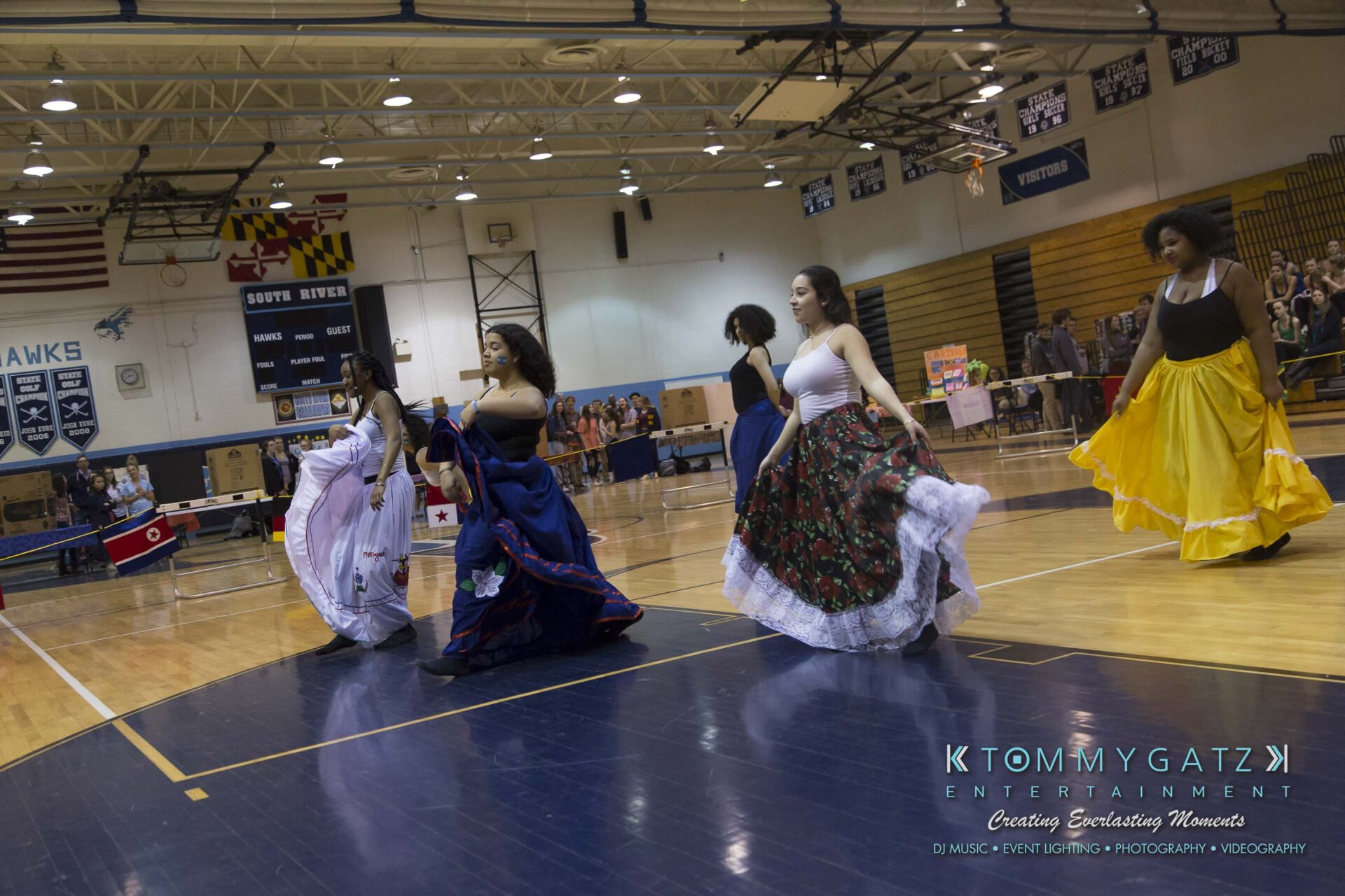 SR-Cultural-Day-2018-Low-Res-WM-20 - TOMMY GATZ ENTERTAINMENT Group of women dancing in colorful skirts in a gymnasium, with flags and audience in the background.