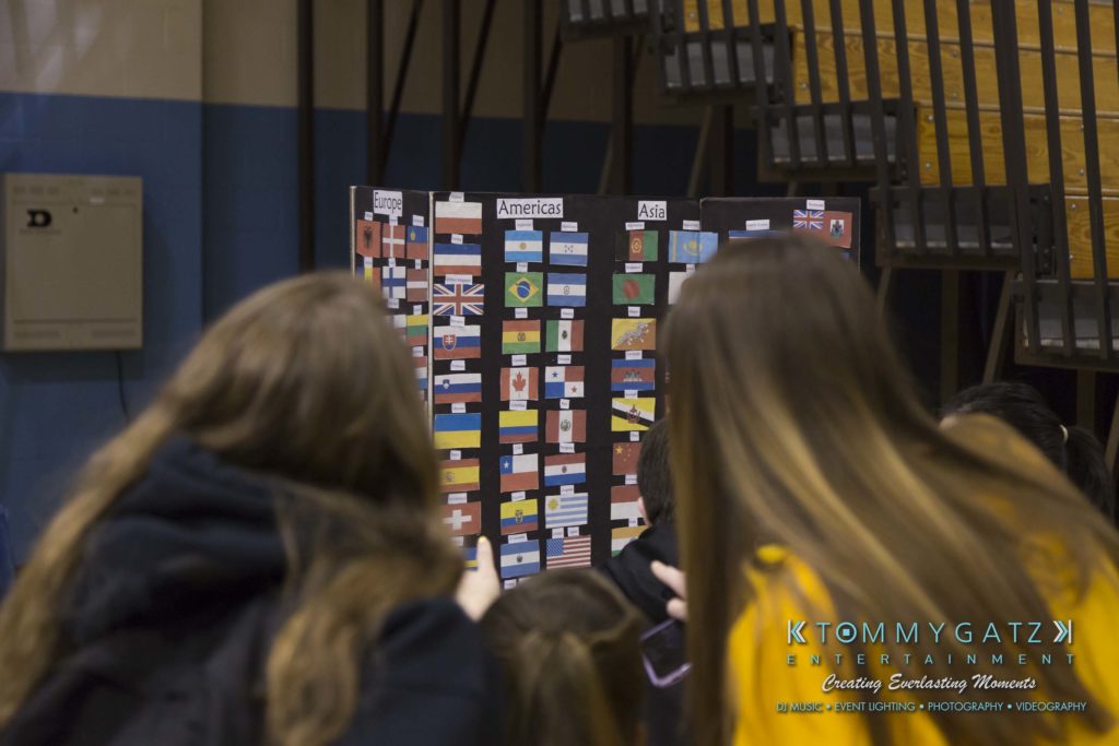 SR-Cultural-Day-2018-Low-Res-WM-29-1024x683 - TOMMY GATZ ENTERTAINMENT People looking at a display board with flags labeled under "America" and "Asia" in a gymnasium setting.
