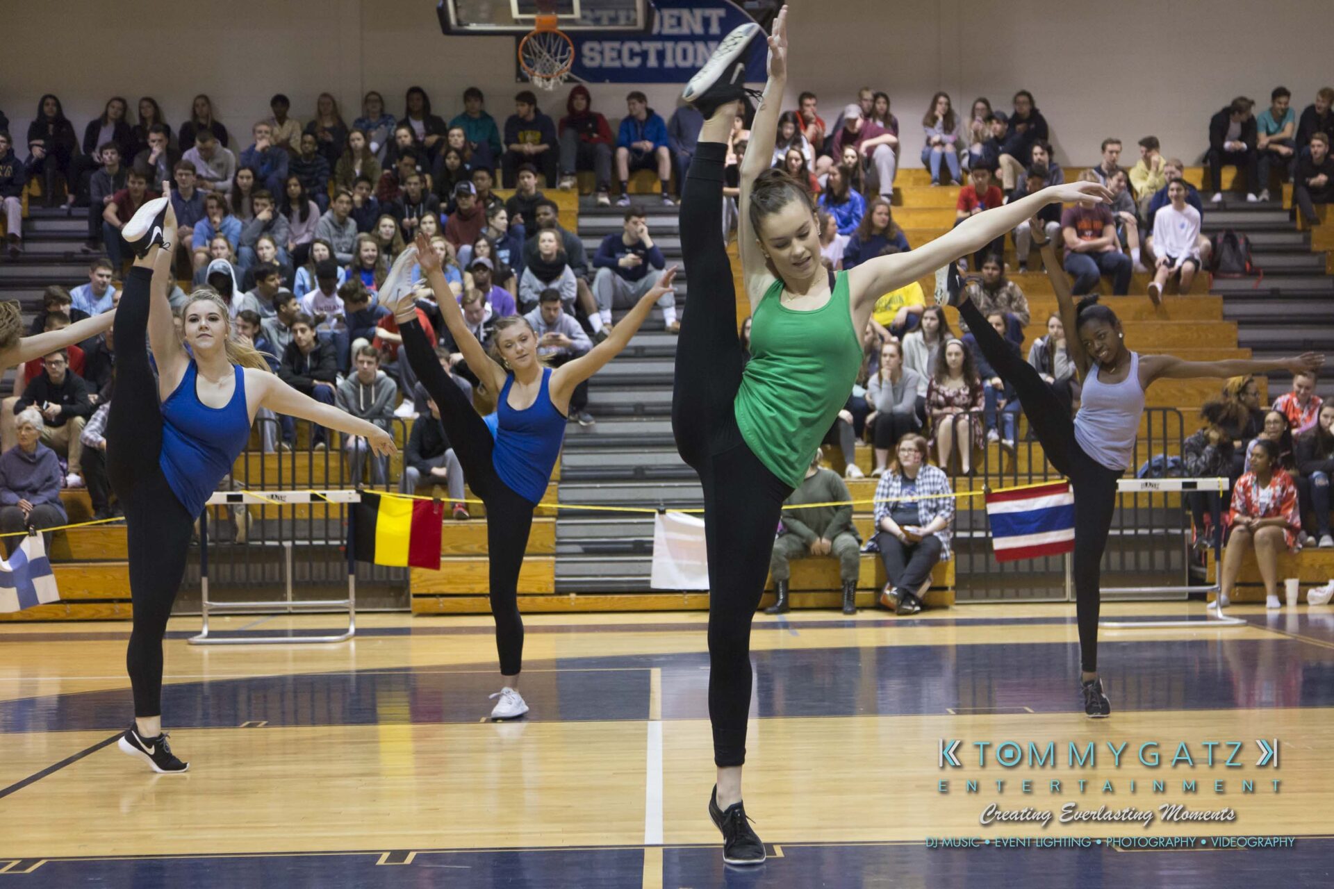 SR-Cultural-Day-2018-Low-Res-WM-33 - TOMMY GATZ ENTERTAINMENT A group of dancers performs a high kick routine on a gym floor, in front of seated spectators. Flags and banners are visible in the background.