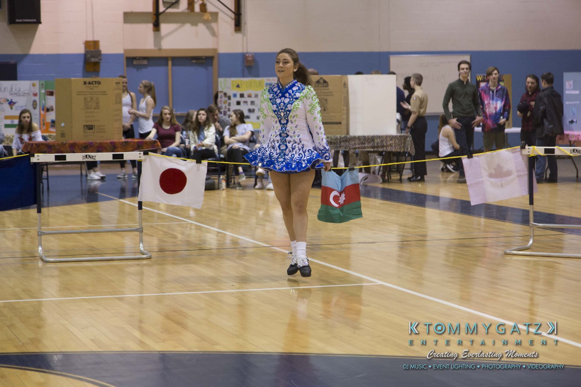 SR-Cultural-Day-2018-Low-Res-WM-35 - TOMMY GATZ ENTERTAINMENT A dancer in a blue and white costume performs indoors. Flags of Japan and Azerbaijan hang nearby. People sit and stand in the background.