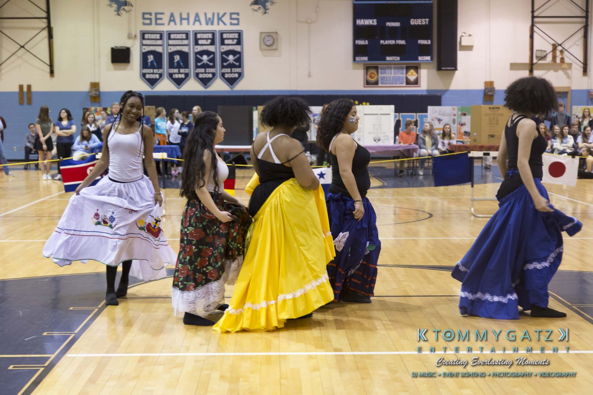 SR-Cultural-Day-2018-Low-Res-WM-43 - TOMMY GATZ ENTERTAINMENT Five women wearing colorful skirts dance in a gymnasium with flags and decorations in the background.