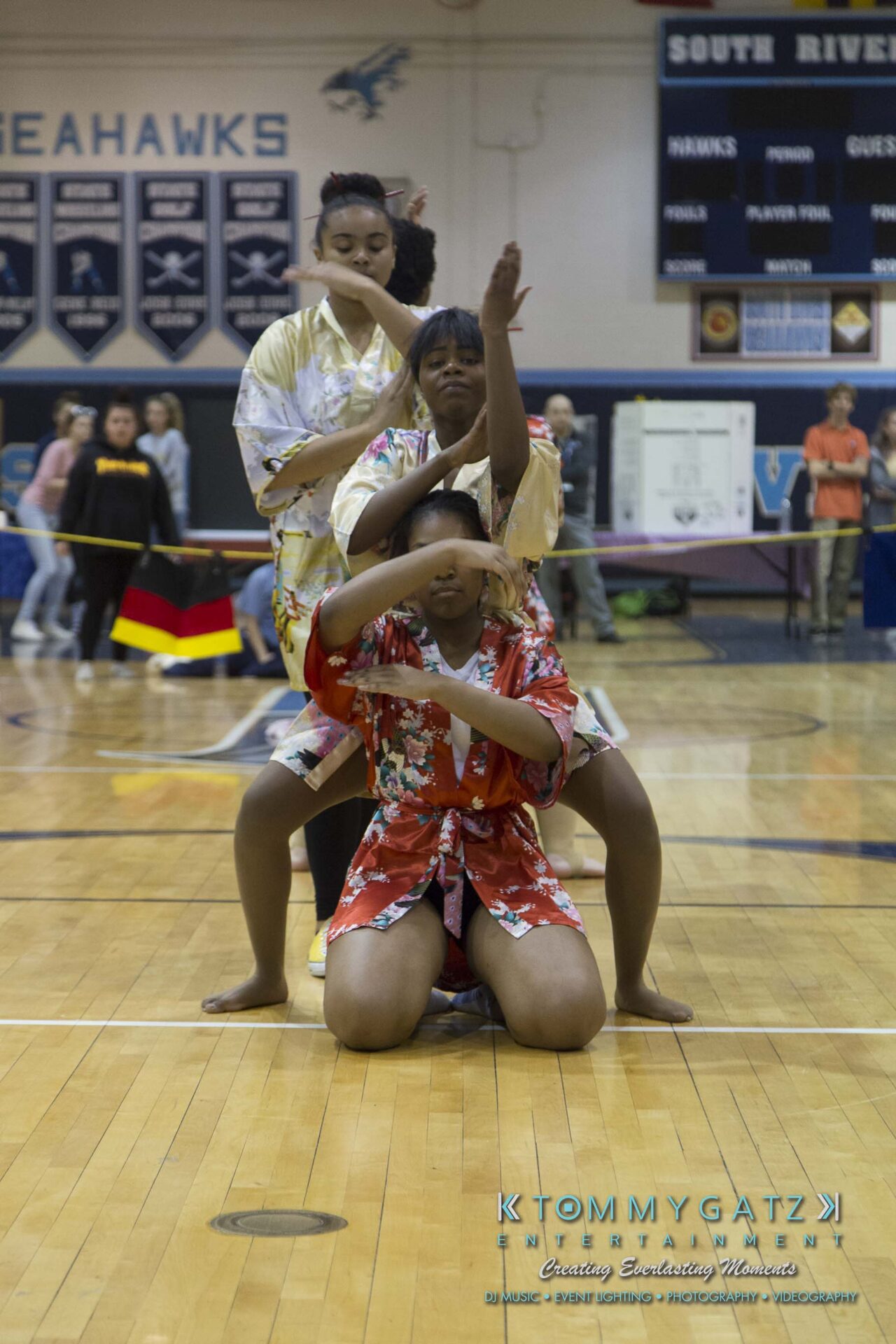 SR-Cultural-Day-2018-Low-Res-WM-55 - TOMMY GATZ ENTERTAINMENT Three people in colorful robes perform a synchronized dance on an indoor basketball court, with spectators and banners in the background.