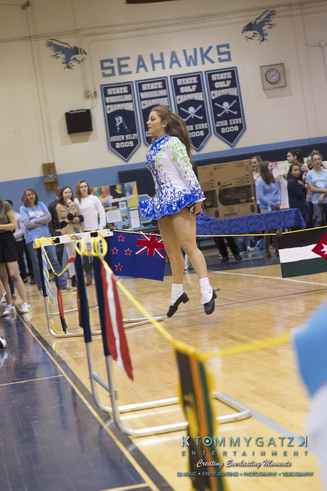SR-Cultural-Day-2018-Low-Res-WM-7 - TOMMY GATZ ENTERTAINMENT A dancer in a blue and green dress performs a jump at an indoor event, surrounded by spectators. Banners with flags are displayed in the background.