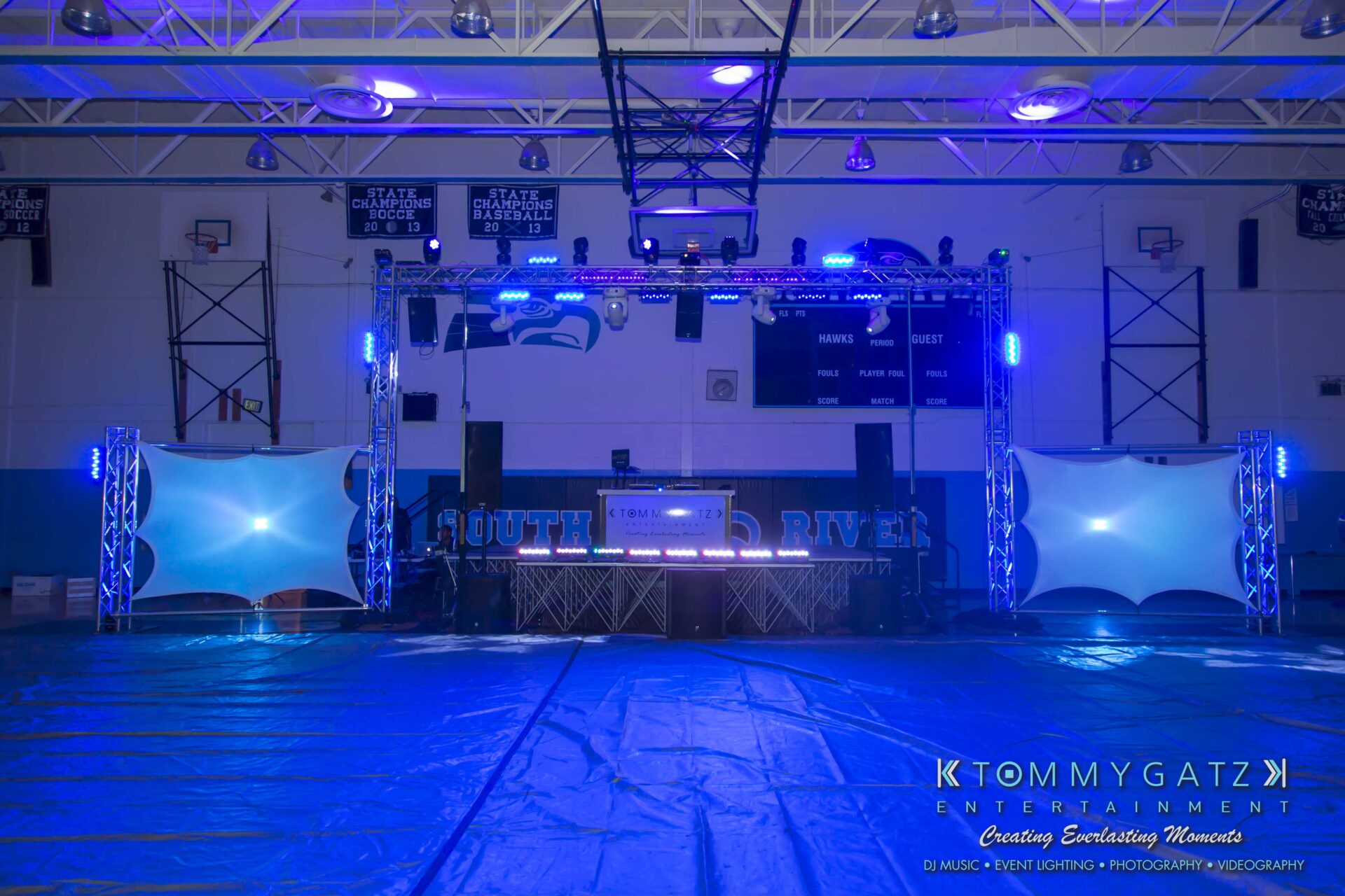 SR-HC-Dance-10717-BLOG-WM-1 1 - TOMMY GATZ ENTERTAINMENT A gymnasium is set up for an event with a DJ booth, blue lighting, and large speakers. A banner reads "South River" on the wall.