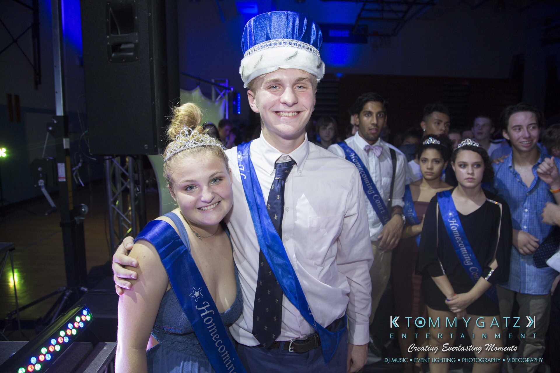 SR-HC-Dance-10717-BLOG-WM-104 - TOMMY GATZ ENTERTAINMENT A young couple wearing sashes and crowns, identified as homecoming royalty, smiles at a school dance event. Other attendees are visible in the background.