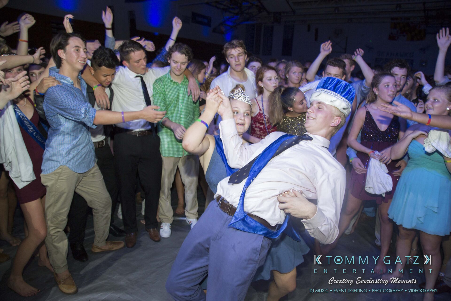 SR-HC-Dance-10717-BLOG-WM-107 - TOMMY GATZ ENTERTAINMENT A group of teenagers dance energetically at a school event. A couple in the center wears blue sashes and crowns.