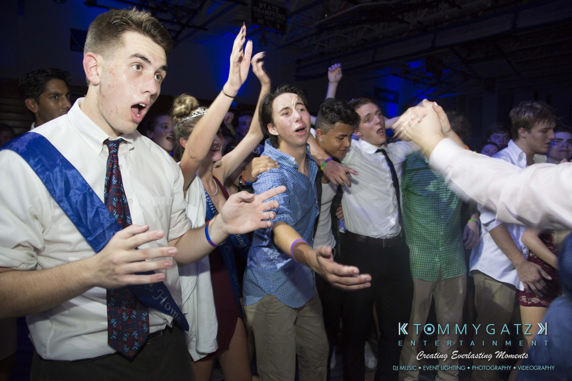 SR-HC-Dance-10717-BLOG-WM-108 - TOMMY GATZ ENTERTAINMENT A group of people in formal attire dance enthusiastically at an indoor event with blue lighting.