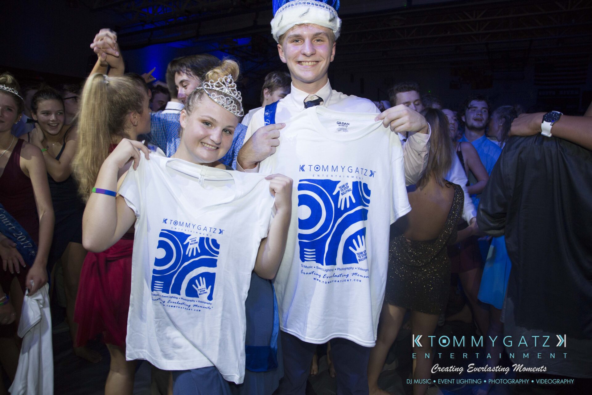 SR-HC-Dance-10717-BLOG-WM-110 - TOMMY GATZ ENTERTAINMENT Two people wearing crowns hold up T-shirts with a logo. They are standing among a group of partygoers in a dimly lit room.