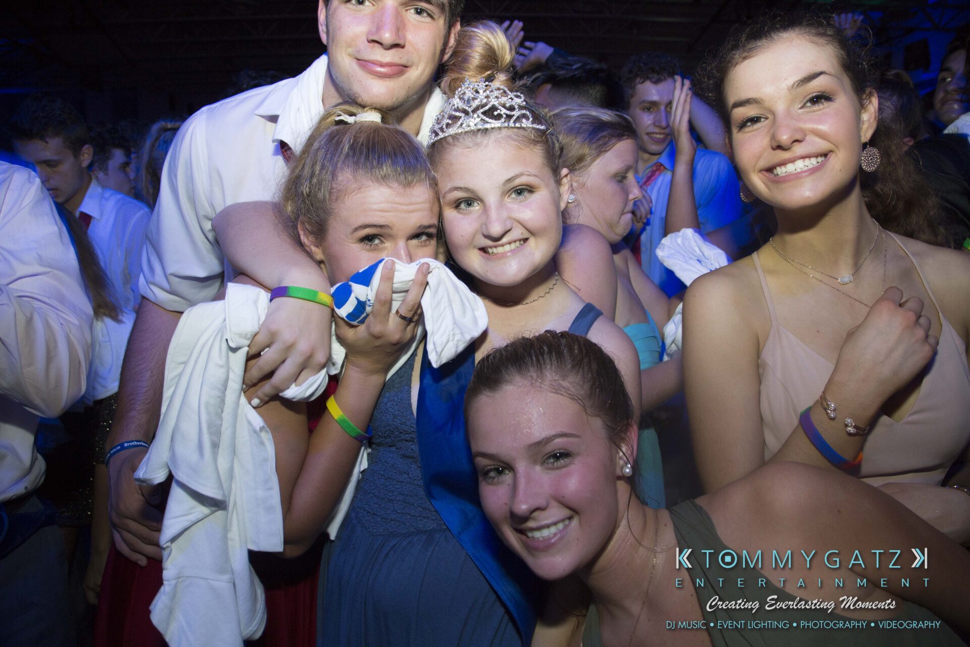 SR-HC-Dance-10717-BLOG-WM-117 - TOMMY GATZ ENTERTAINMENT A group of young people in formal attire smile and pose for a photo at an indoor event. Some wear wristbands and hold cloth items. A person in the center wears a tiara.