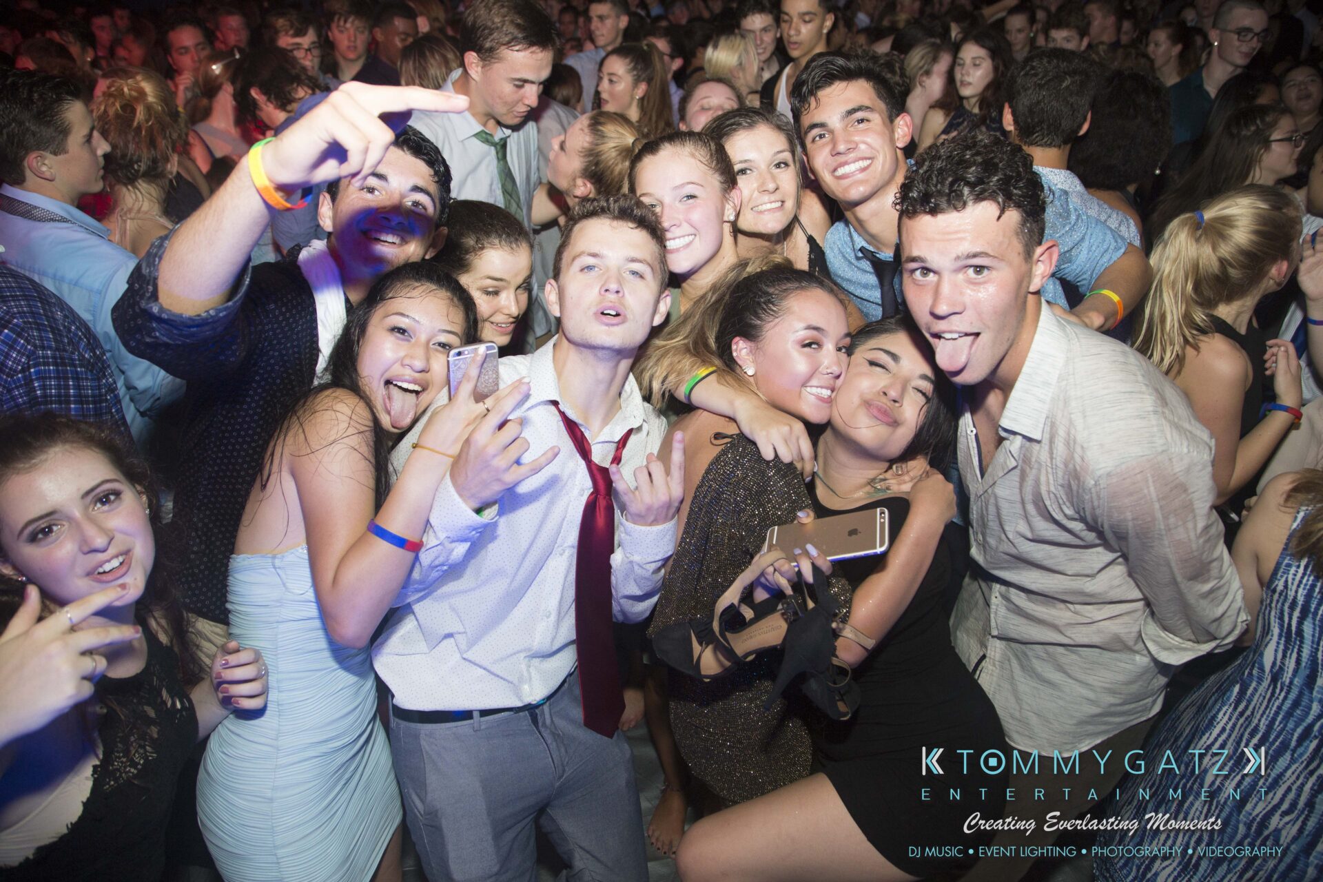 SR-HC-Dance-10717-BLOG-WM-144 - TOMMY GATZ ENTERTAINMENT Group of people posing in a crowded party setting, many smiling and taking selfies, with colorful lighting in the background.
