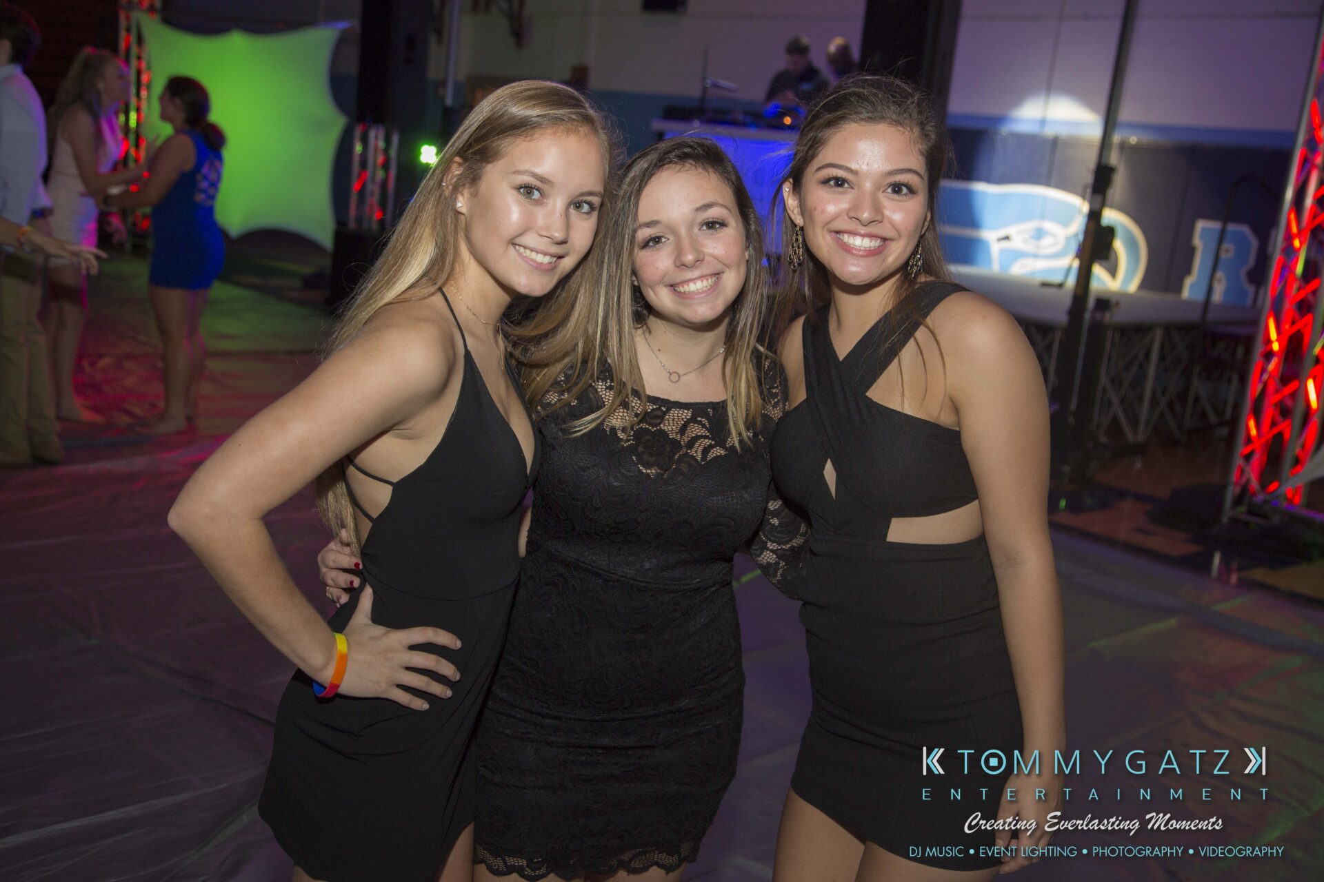 SR-HC-Dance-10717-BLOG-WM-18 - TOMMY GATZ ENTERTAINMENT Three people in black dresses pose closely together at an indoor event, smiling at the camera.