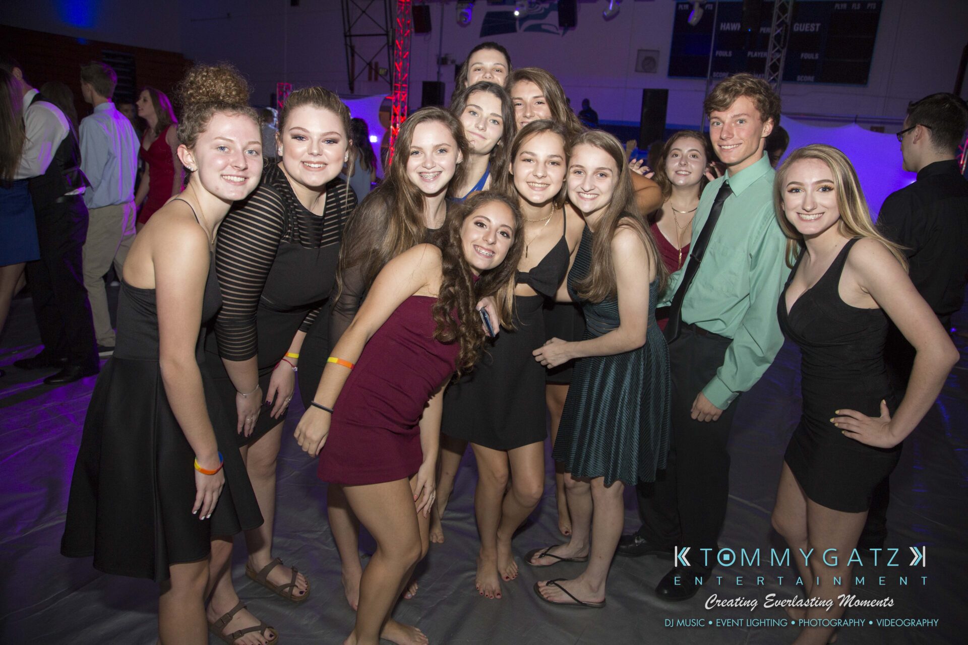 SR-HC-Dance-10717-BLOG-WM-24 - TOMMY GATZ ENTERTAINMENT A group of young people in formal attire pose together on a dance floor, with colorful lights in the background.