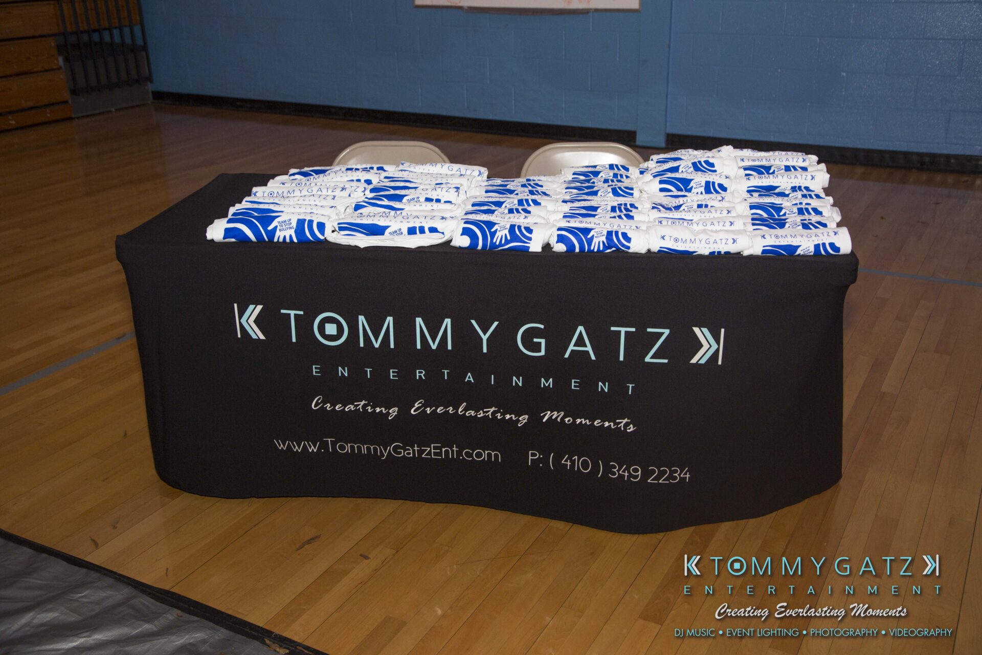 SR-HC-Dance-10717-BLOG-WM-25 - TOMMY GATZ ENTERTAINMENT A table with a black tablecloth displaying "Tommy Gatz Entertainment" is lined with stacked blue and white towels, in a gym setting.