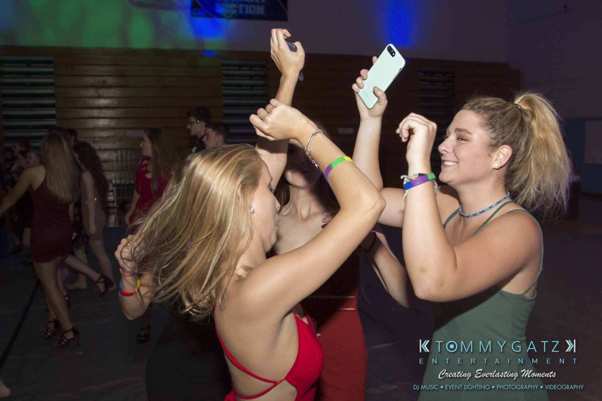 SR-HC-Dance-10717-BLOG-WM-33 - TOMMY GATZ ENTERTAINMENT People dancing energetically at a party, with some holding up a smartphone to capture the moment. The background features dim lighting and a few people in the distance.