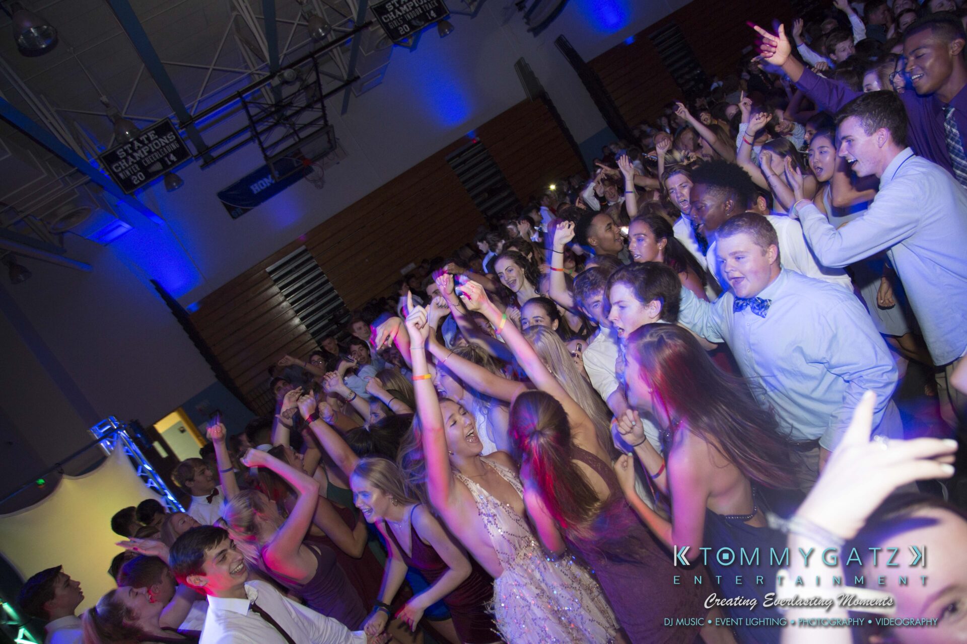 SR-HC-Dance-10717-BLOG-WM-42 - TOMMY GATZ ENTERTAINMENT A large group of young people dancing energetically at an indoor event with colorful lighting and a DJ booth visible.