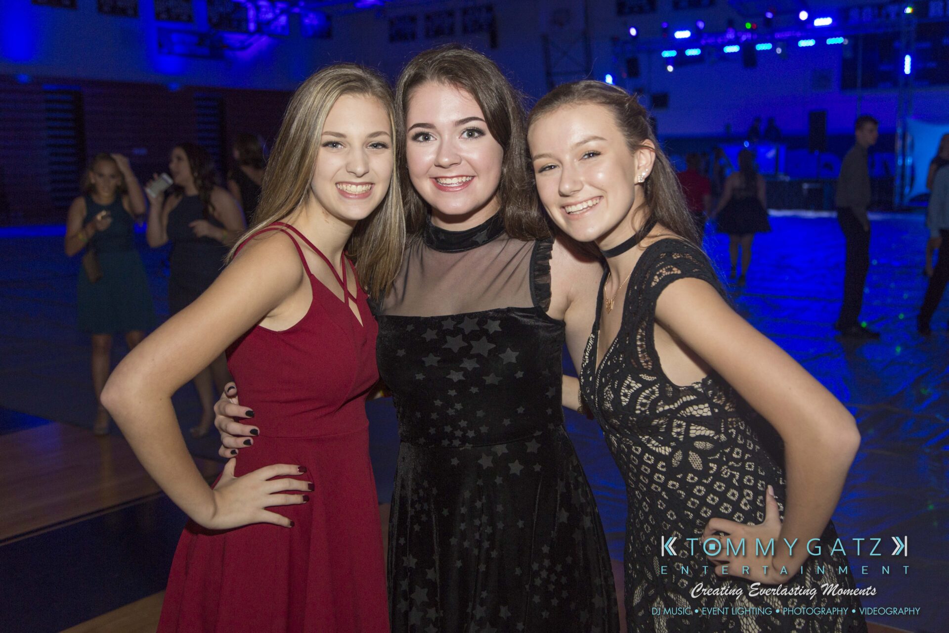 SR-HC-Dance-10717-BLOG-WM-5 1 - TOMMY GATZ ENTERTAINMENT Three young women in dresses smile at the camera in a dimly lit event space with blue lighting.
