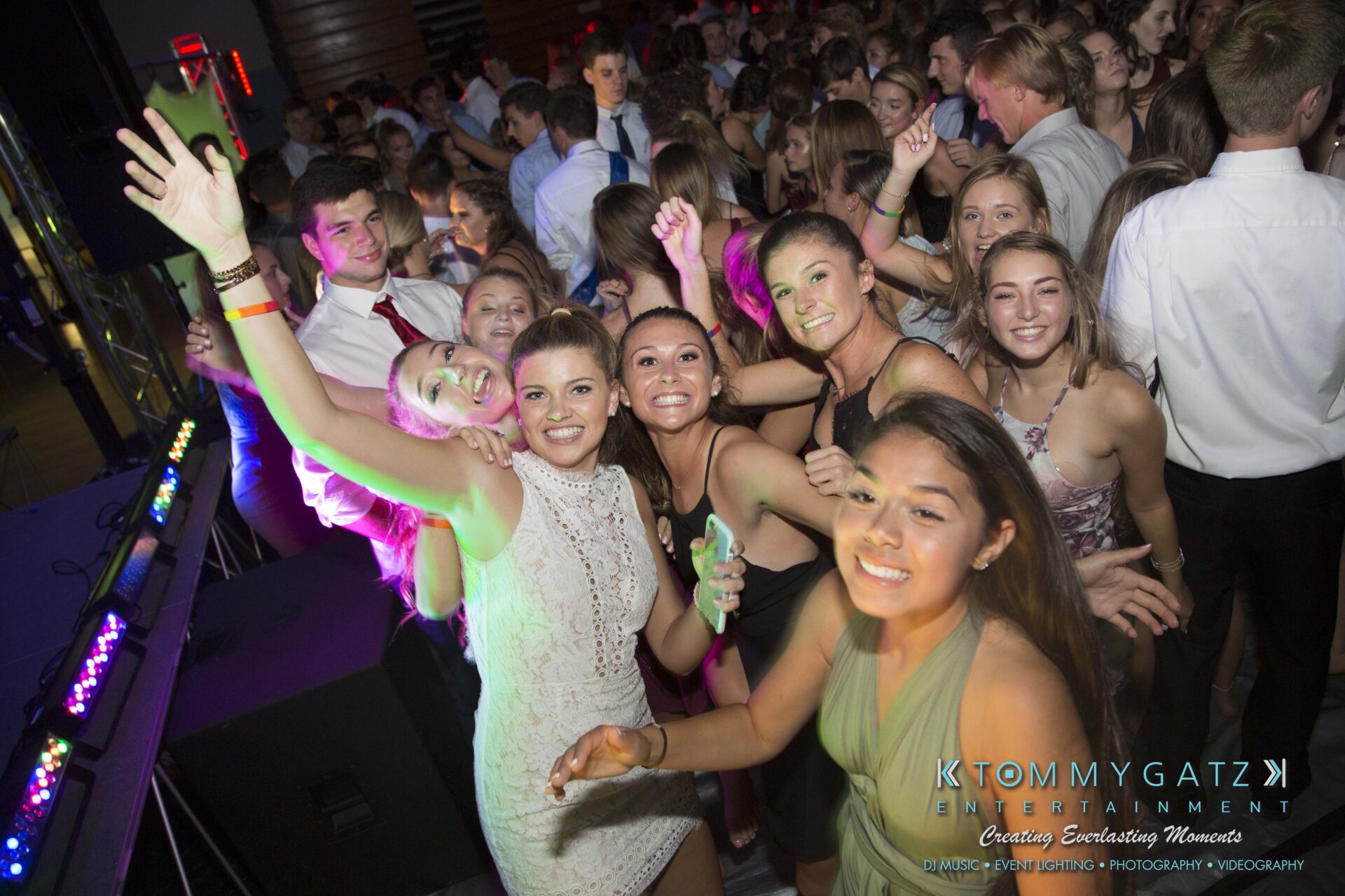 SR-HC-Dance-10717-BLOG-WM-51 - TOMMY GATZ ENTERTAINMENT A group of young people, dressed in formal attire, are gathered closely together at a lively event, some smiling at the camera. Colorful lights illuminate the scene.