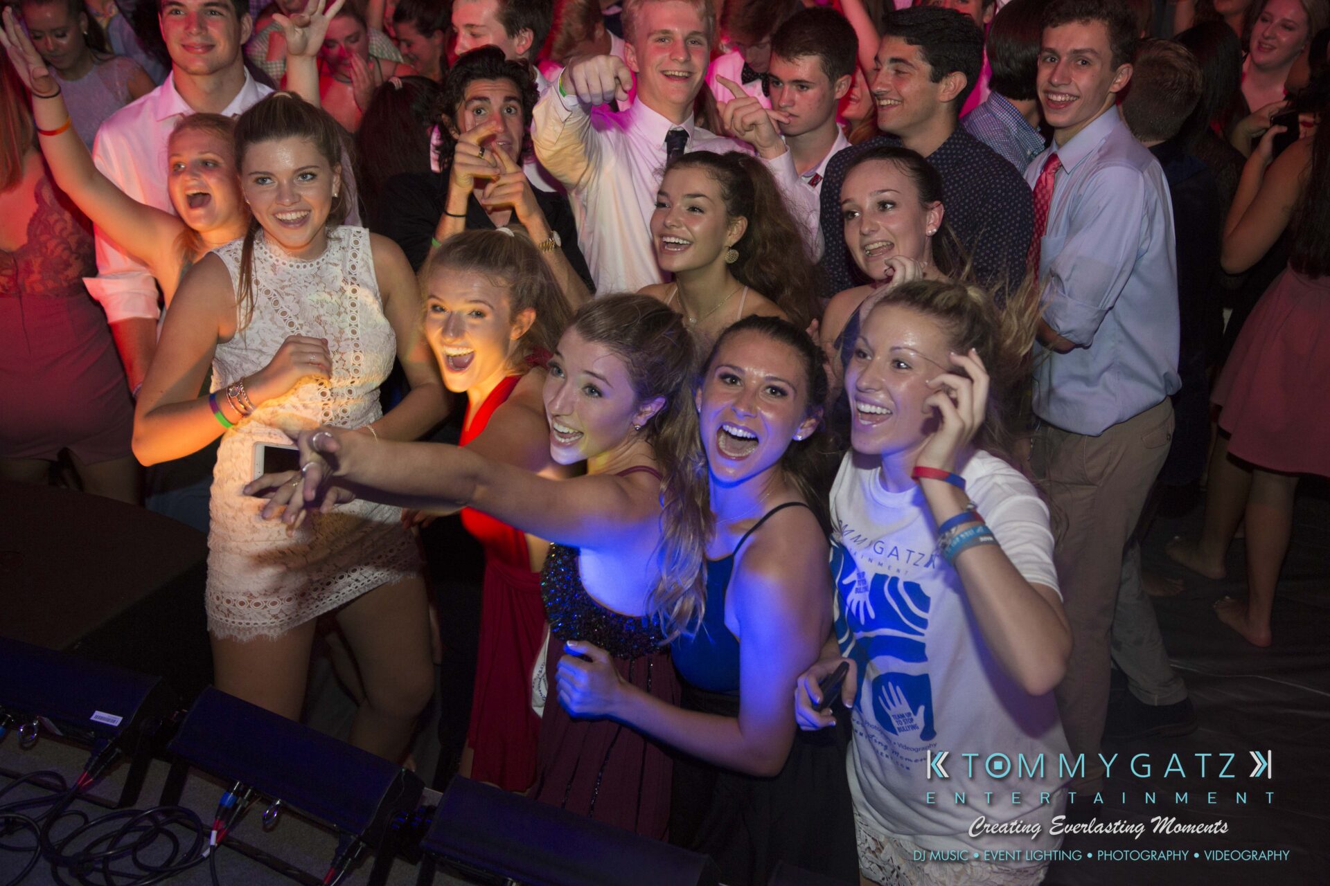 SR-HC-Dance-10717-BLOG-WM-65 - TOMMY GATZ ENTERTAINMENT A group of young people at a party, smiling and taking a selfie. They're dressed in casual and semi-formal attire. The background features colorful lighting and a crowded dance floor.