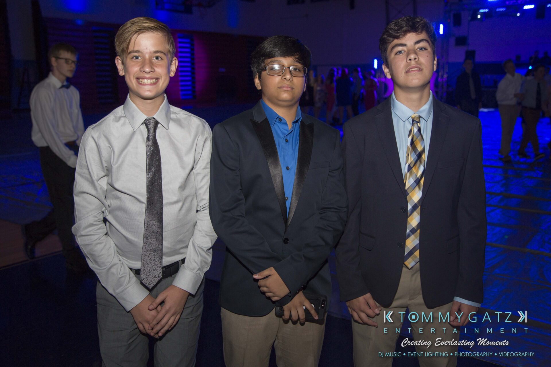 SR-HC-Dance-10717-BLOG-WM-7 - TOMMY GATZ ENTERTAINMENT Three young individuals in formal attire pose for a photo in a dimly lit event space with blue lighting.