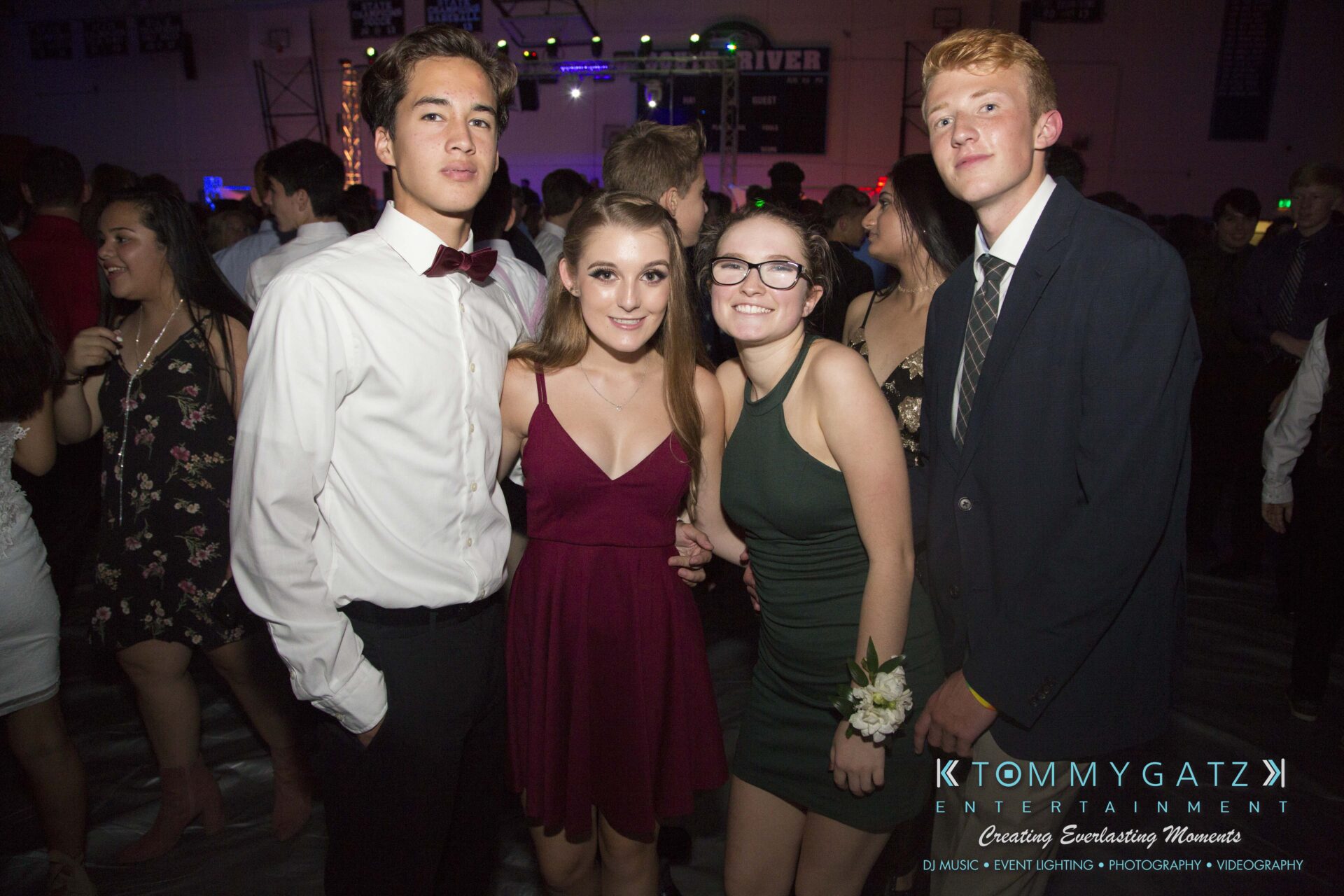 SR-HC-Dance-10717-BLOG-WM-73 - TOMMY GATZ ENTERTAINMENT Four teenagers posing together at a formal event; three are wearing dresses, and one is in a suit. The setting appears to be a party or a dance with other attendees in the background.