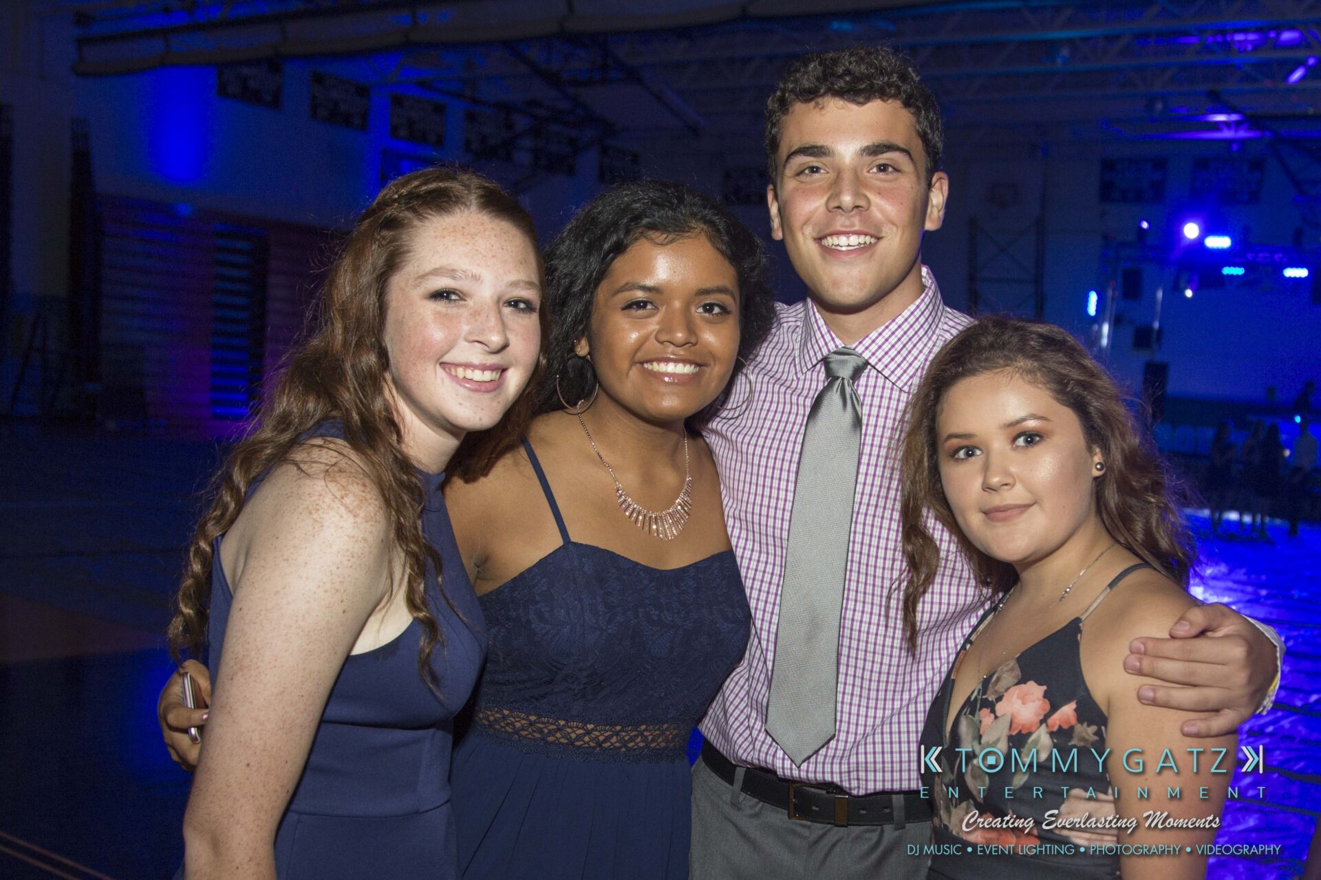 SR-HC-Dance-10717-BLOG-WM-8 - TOMMY GATZ ENTERTAINMENT Four people smiling and posing together at an indoor event with blue lighting.