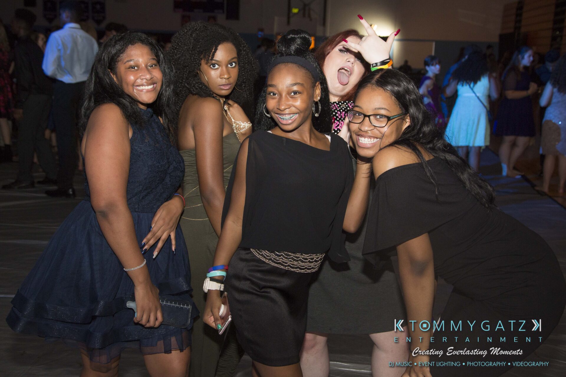 SR-HC-Dance-10717-BLOG-WM-84 - TOMMY GATZ ENTERTAINMENT A group of five people posing and smiling at a lively indoor event, with colorful lighting and decorations in the background.