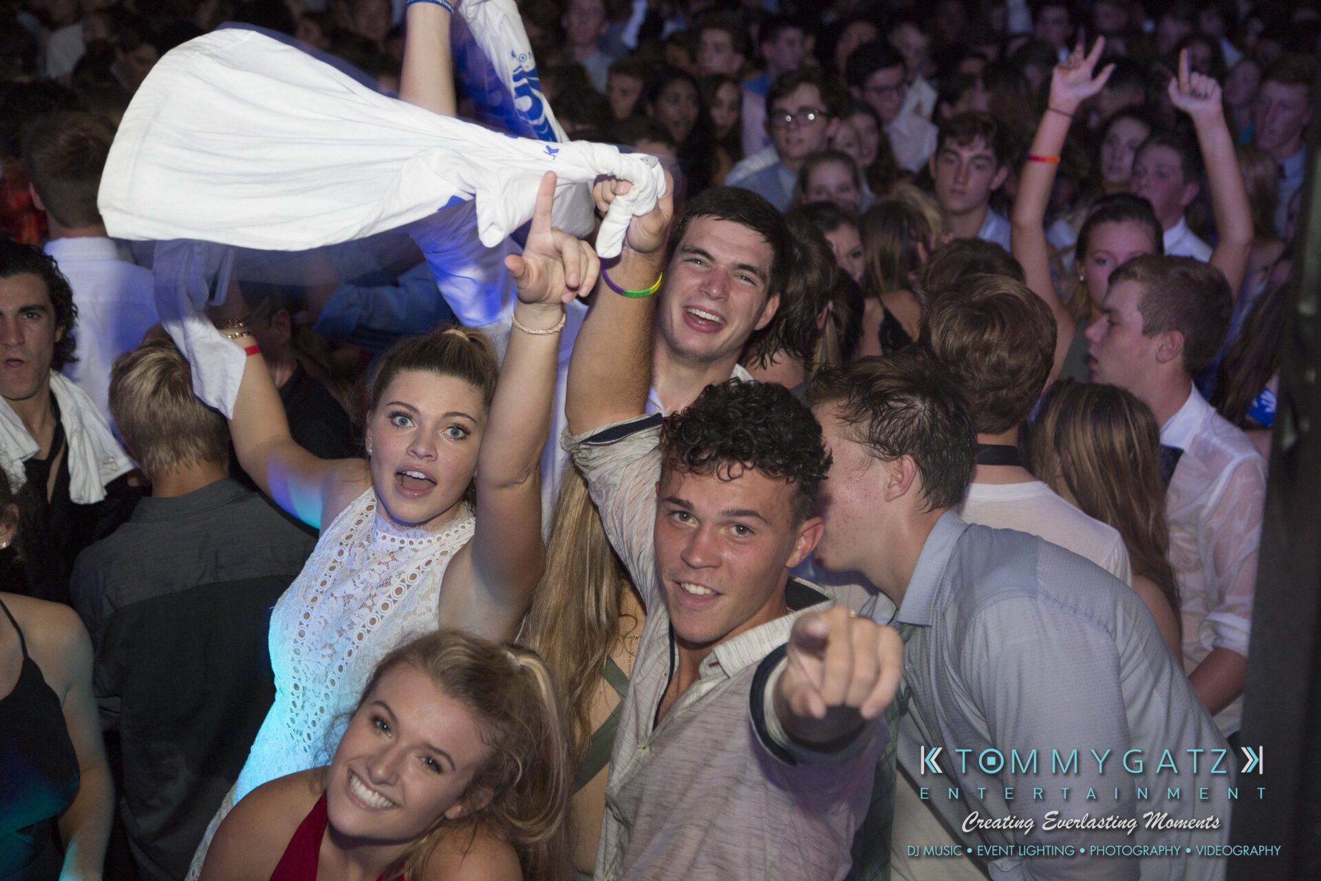 SR-HC-Dance-10717-BLOG-WM-87 - TOMMY GATZ ENTERTAINMENT A group of people are dancing excitedly in a crowded venue, some raising their hands or pointing at the camera, with colorful lighting in the background.