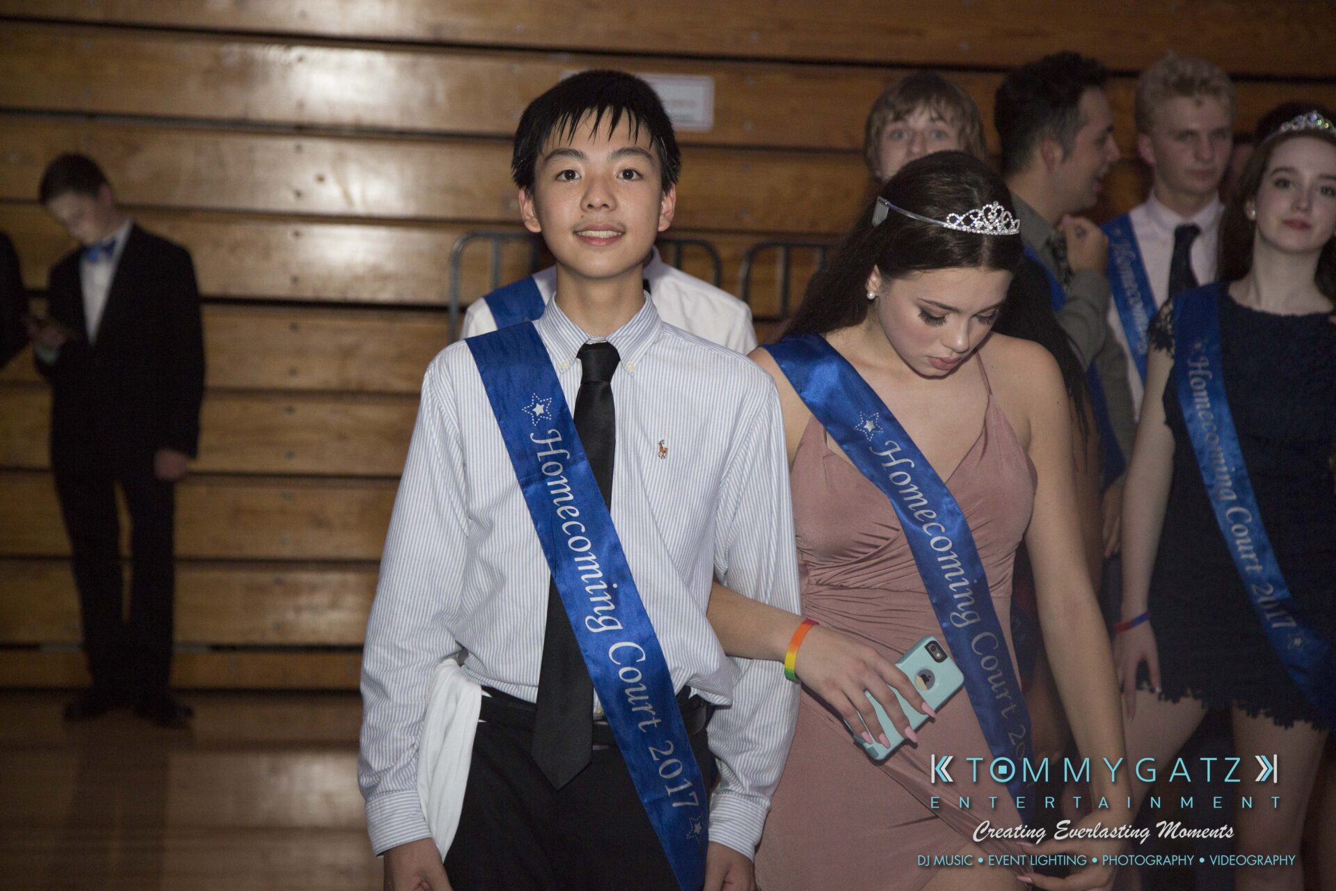 SR-HC-Dance-10717-BLOG-WM-89 - TOMMY GATZ ENTERTAINMENT A group of people wearing "Homecoming Court 2017" sashes stands together in a dimly lit gymnasium.