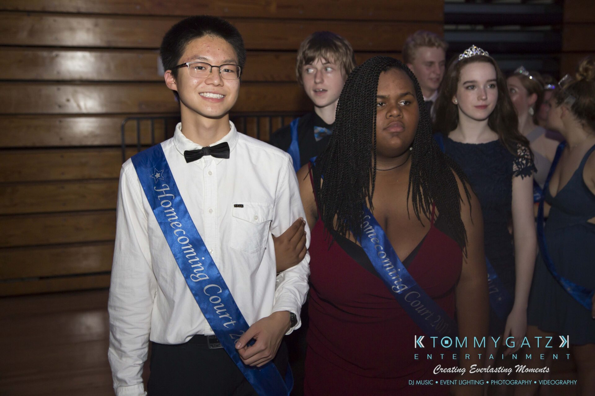 SR-HC-Dance-10717-BLOG-WM-90 - TOMMY GATZ ENTERTAINMENT A group of students in formal attire, with two wearing "Homecoming Court" sashes, stand in a dimly lit room.