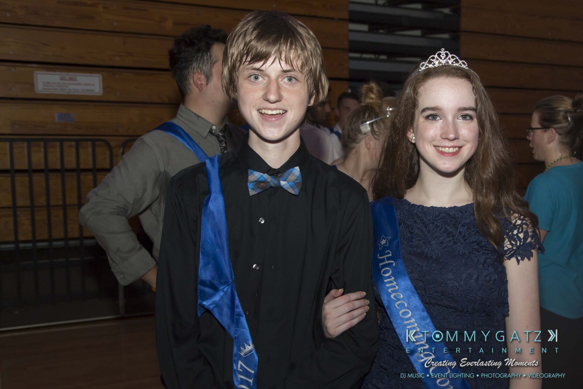 SR-HC-Dance-10717-BLOG-WM-91 - TOMMY GATZ ENTERTAINMENT Two young people wearing blue sashes and formal attire pose arm-in-arm at a dance event. Others are visible in the background.