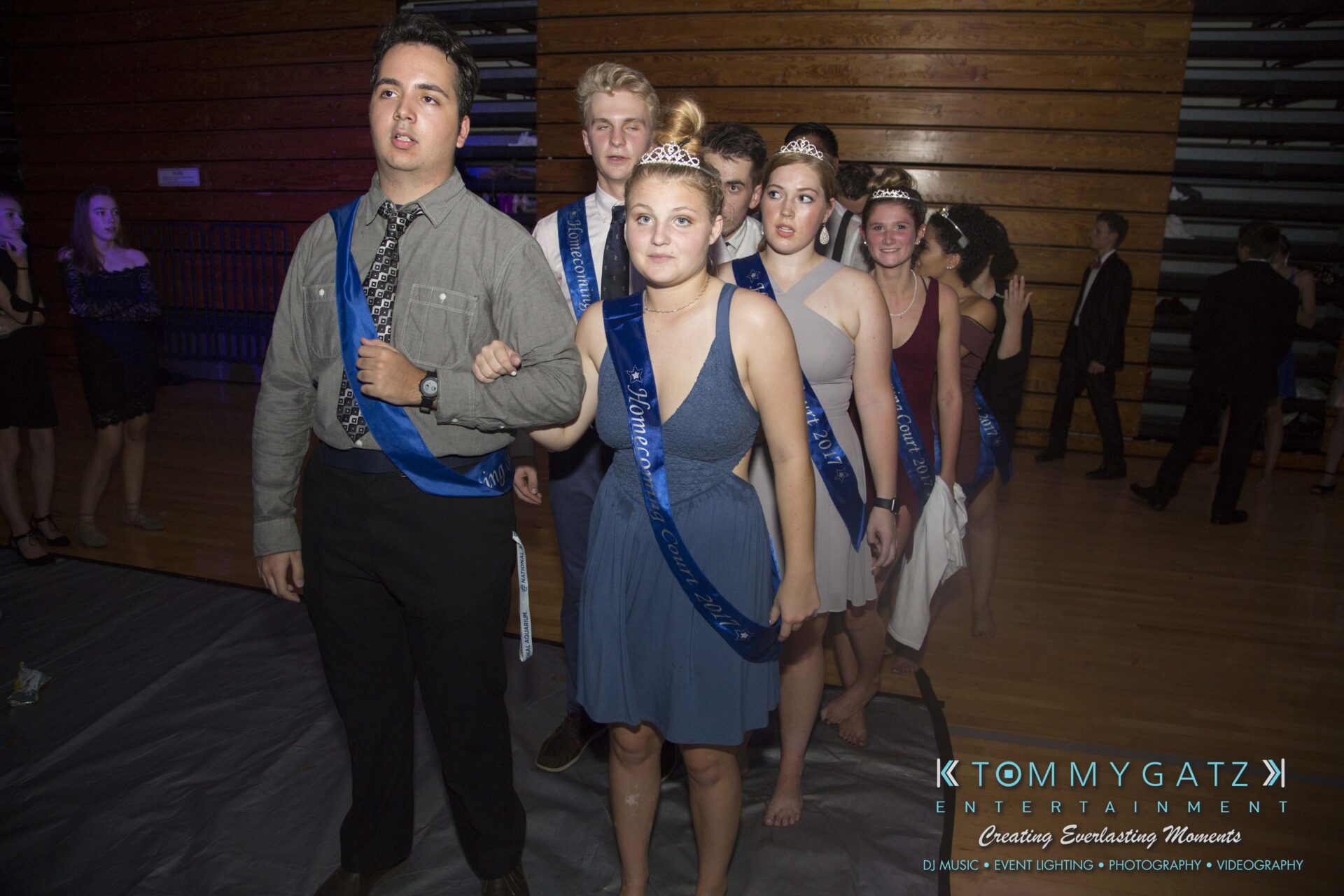 SR-HC-Dance-10717-BLOG-WM-93 - TOMMY GATZ ENTERTAINMENT A group of young people in formal attire and tiaras line up in a gymnasium, holding blue sashes.
