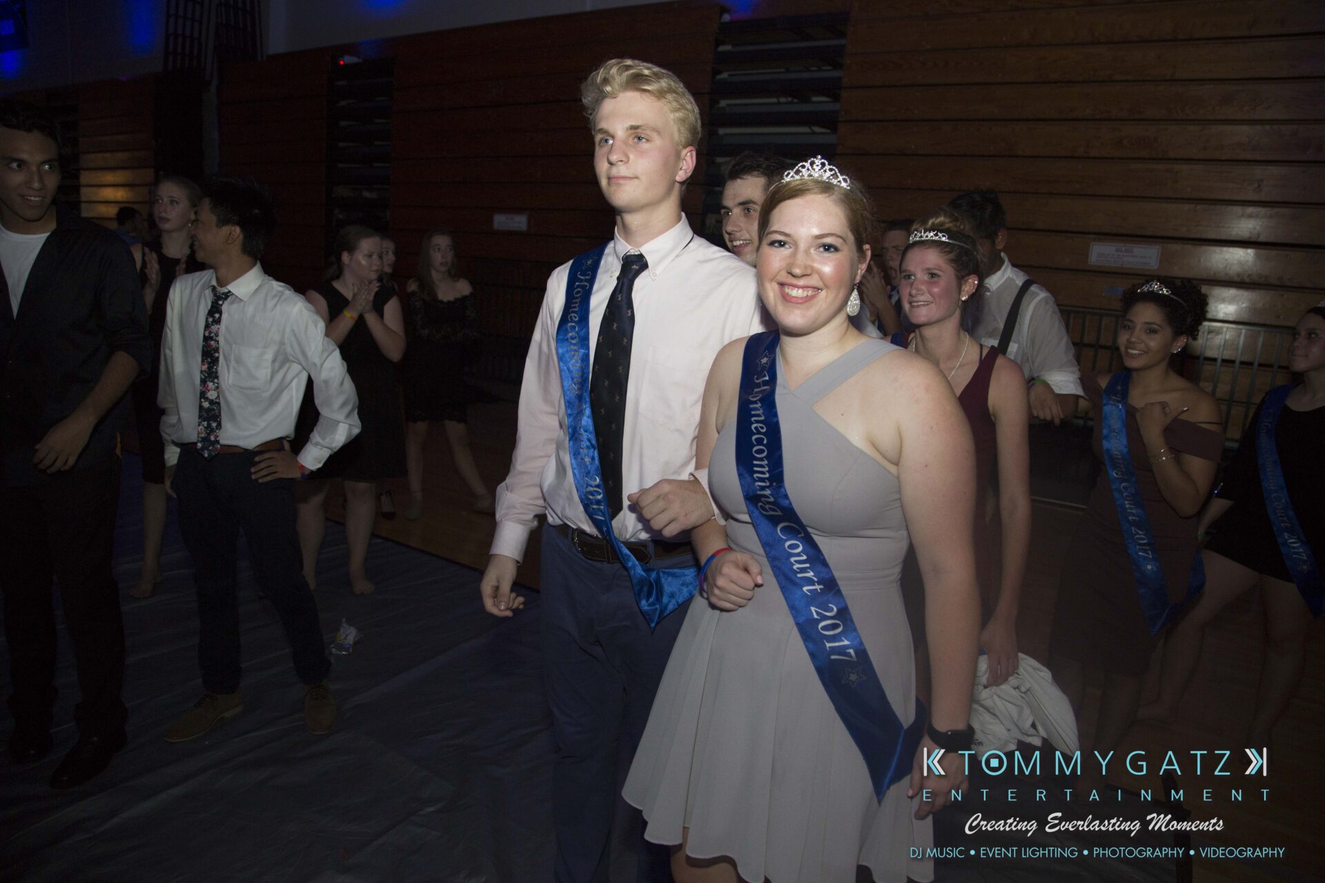 SR-HC-Dance-10717-BLOG-WM-95 - TOMMY GATZ ENTERTAINMENT A young woman in a tiara and sash stands next to a young man in a sash. They're in a gym with people in the background, dressed for an event.