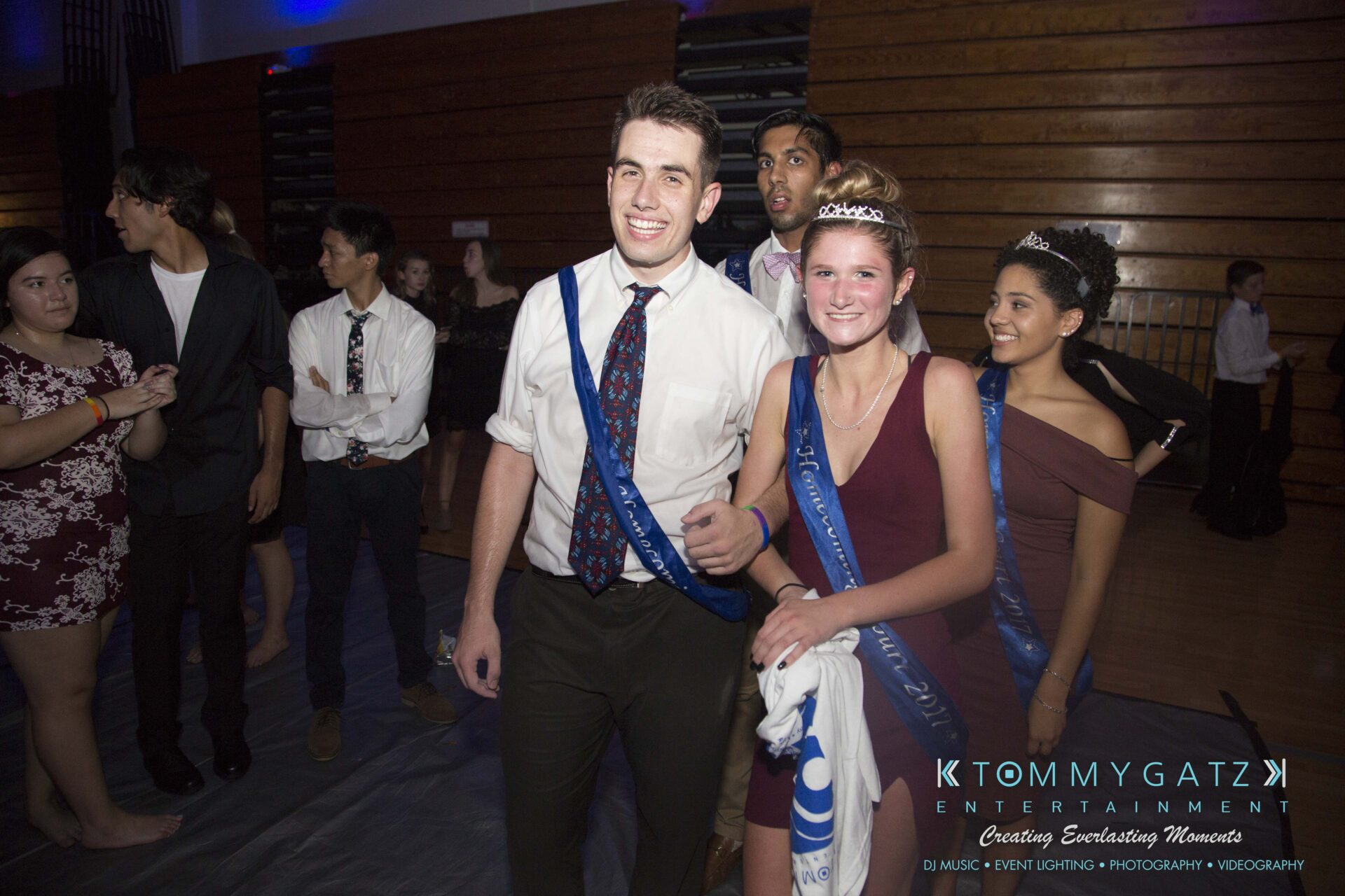 SR-HC-Dance-10717-BLOG-WM-96 - TOMMY GATZ ENTERTAINMENT A group of young adults in formal attire, some wearing sashes and crowns, pose for a photo at an indoor event with a wooden backdrop.