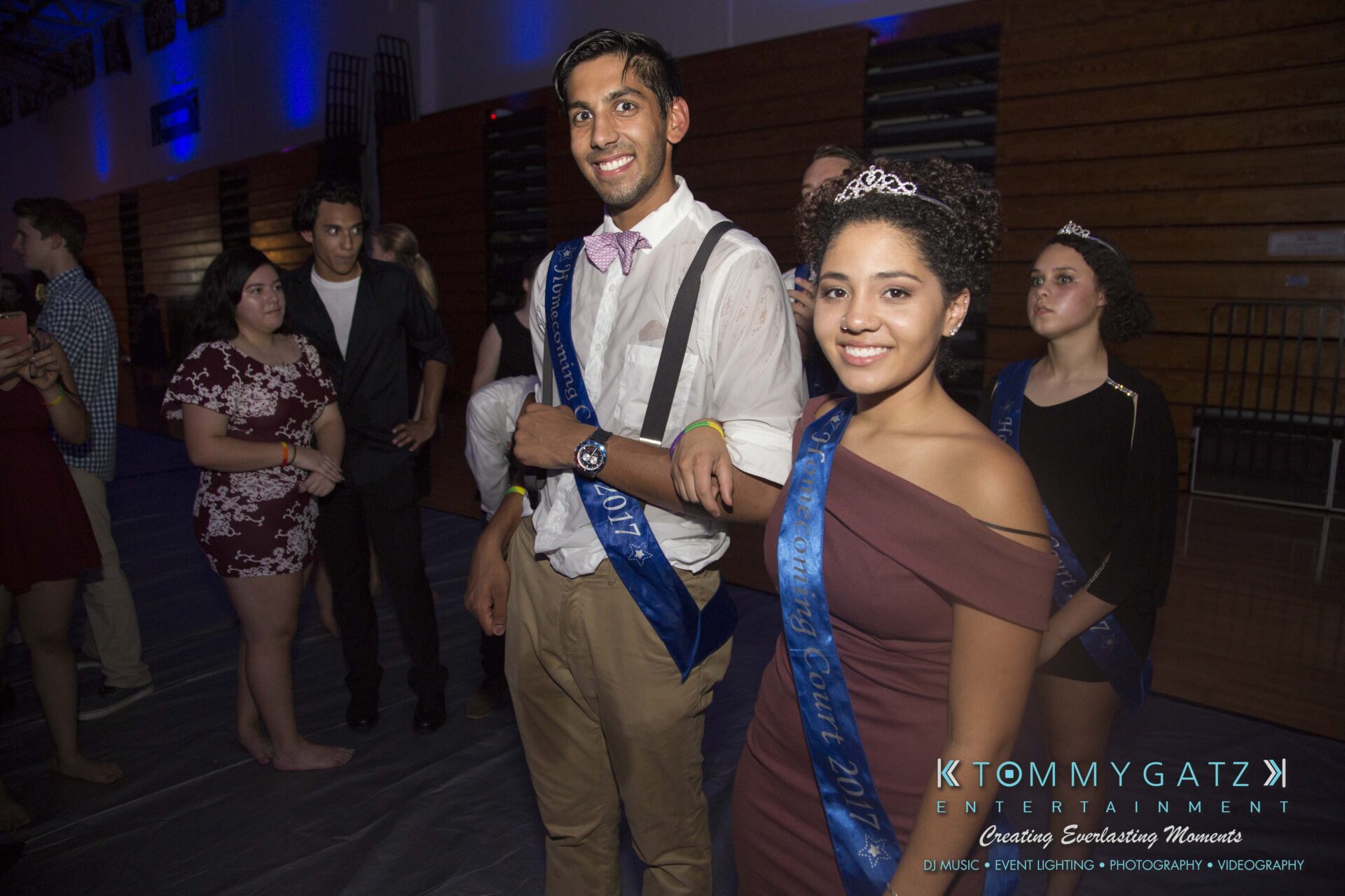 SR-HC-Dance-10717-BLOG-WM-97 - TOMMY GATZ ENTERTAINMENT A group of people, some wearing sashes and tiaras, stand indoors in a gymnasium with blue lighting.