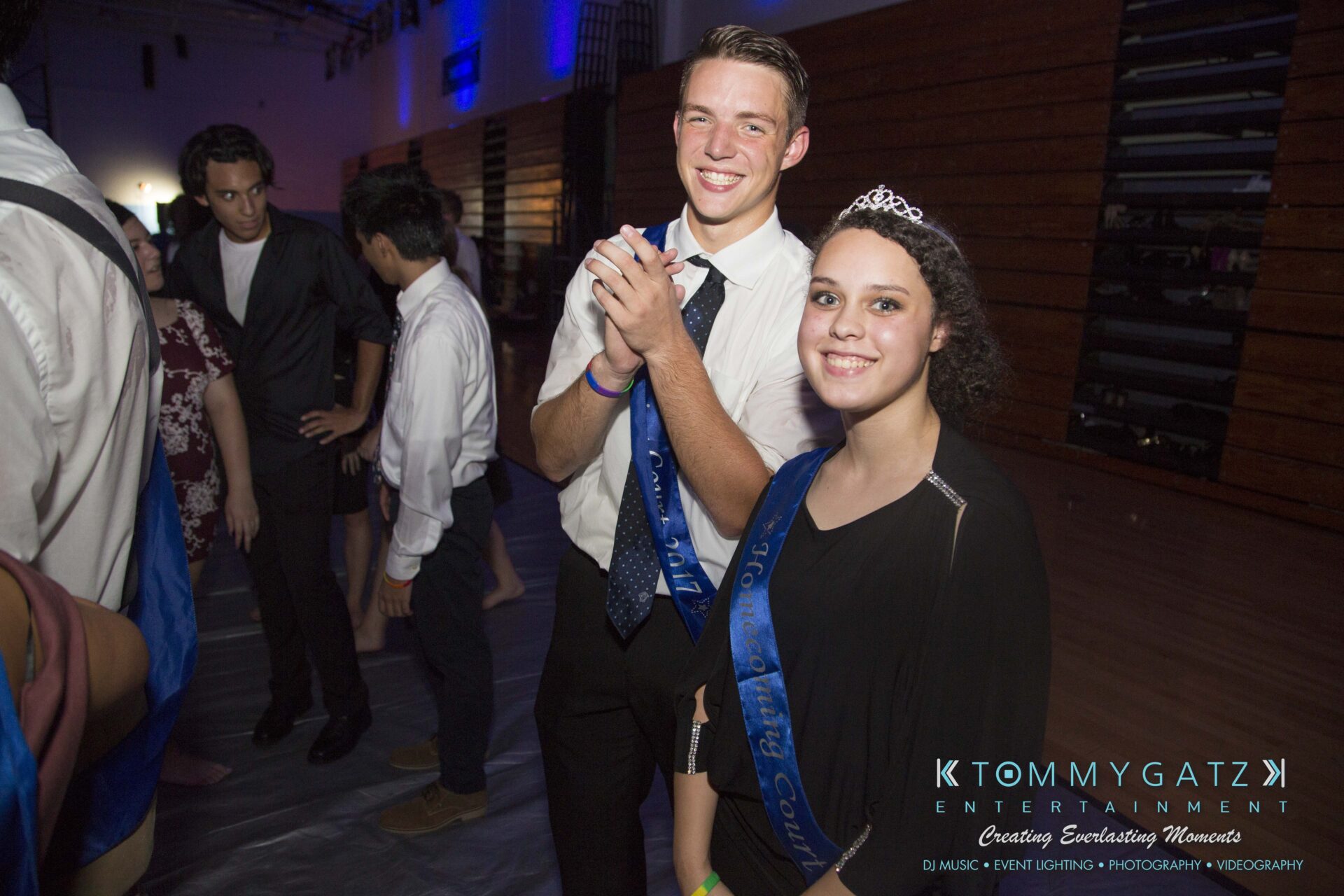 SR-HC-Dance-10717-BLOG-WM-98 - TOMMY GATZ ENTERTAINMENT A young man and woman wearing sashes stand together, smiling, at an indoor event.