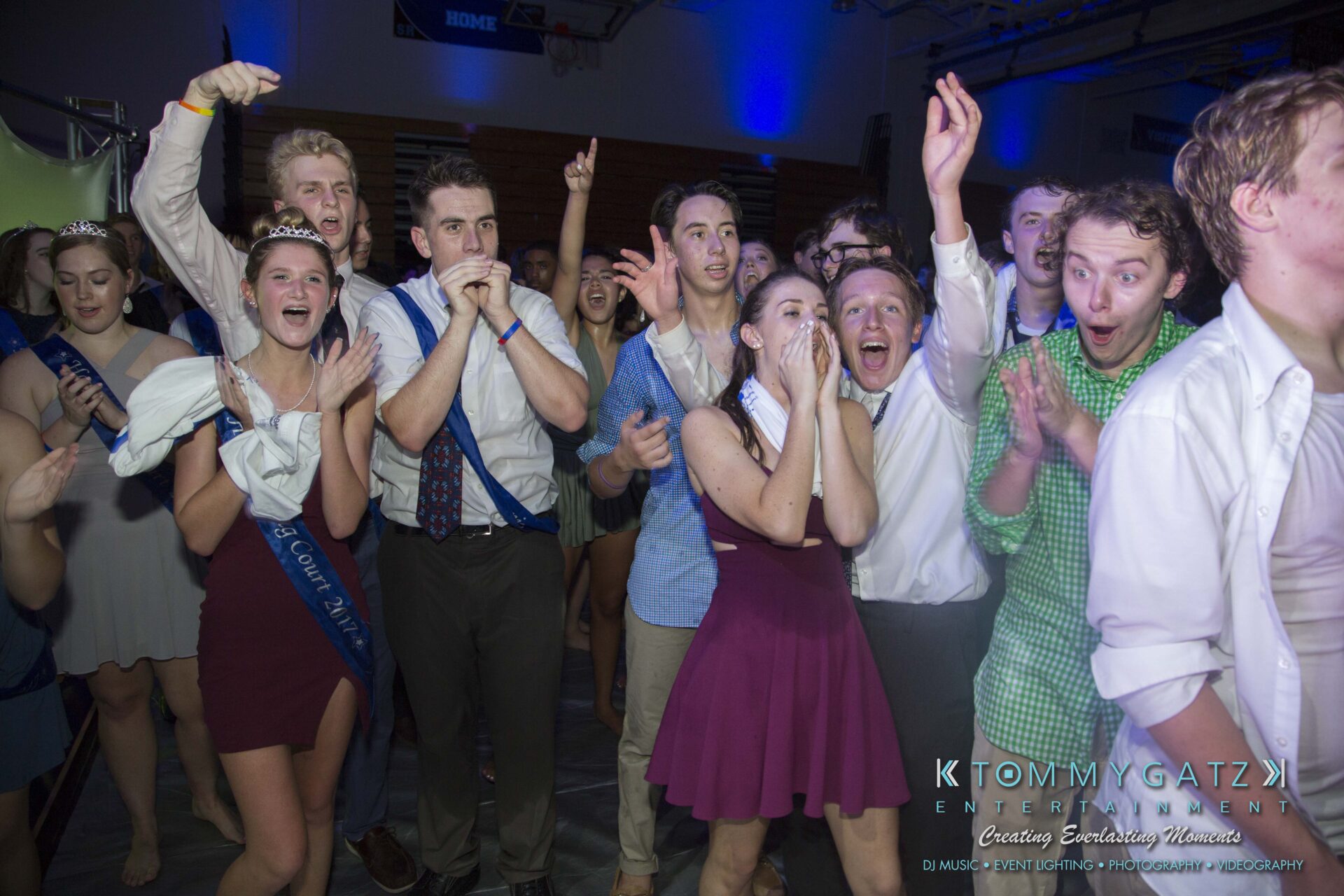 SR-HC-Dance-10717-BLOG-WM-99 - TOMMY GATZ ENTERTAINMENT A group of people at a party reacting excitedly, some with hands raised or on their faces, in a dimly lit room with blue lights.
