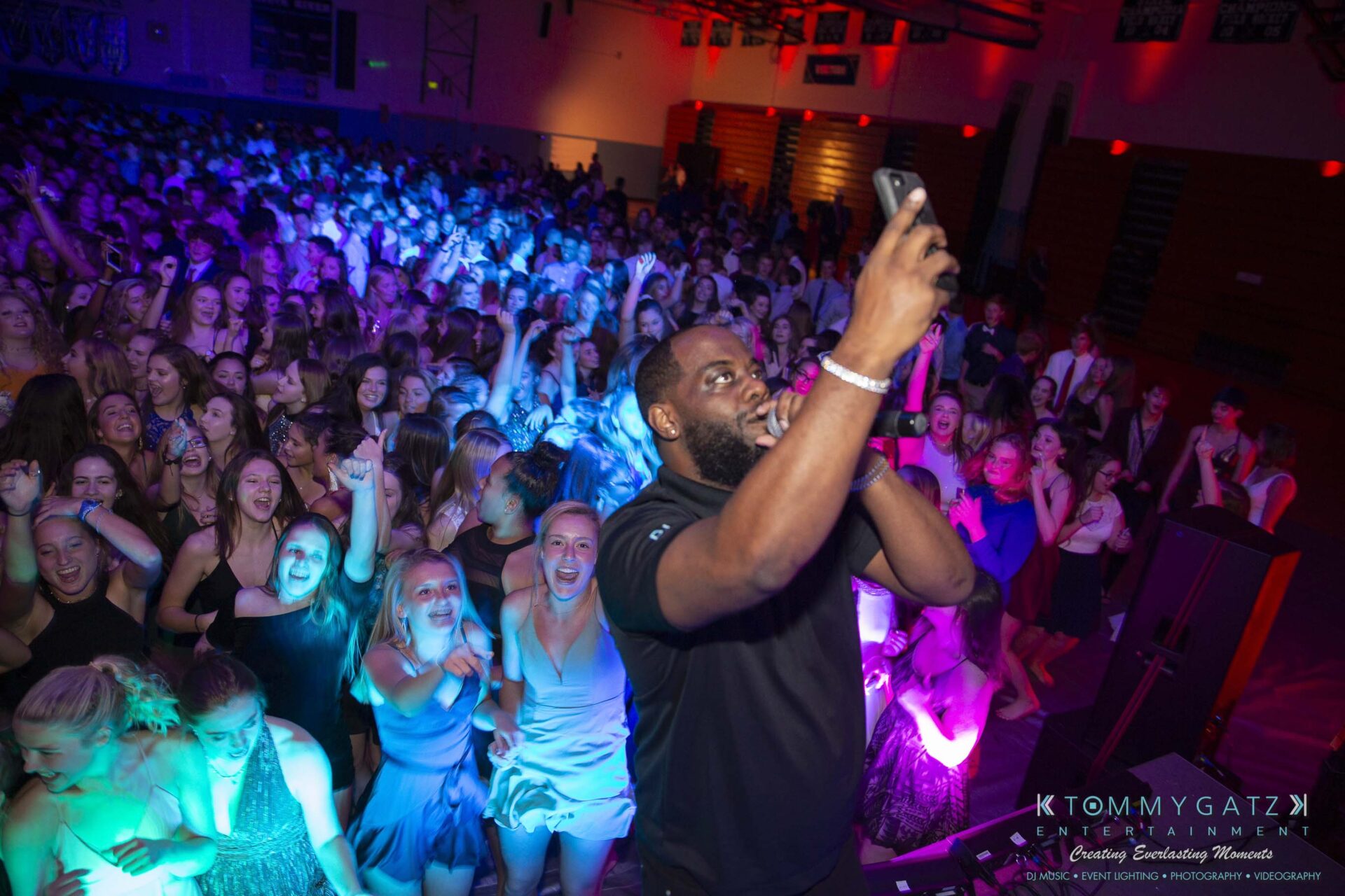 SR-Homecoming-102718-Carl-Low-Res-22 - TOMMY GATZ ENTERTAINMENT A man takes a selfie on stage in front of a large, cheering crowd at an indoor event with colorful lights.