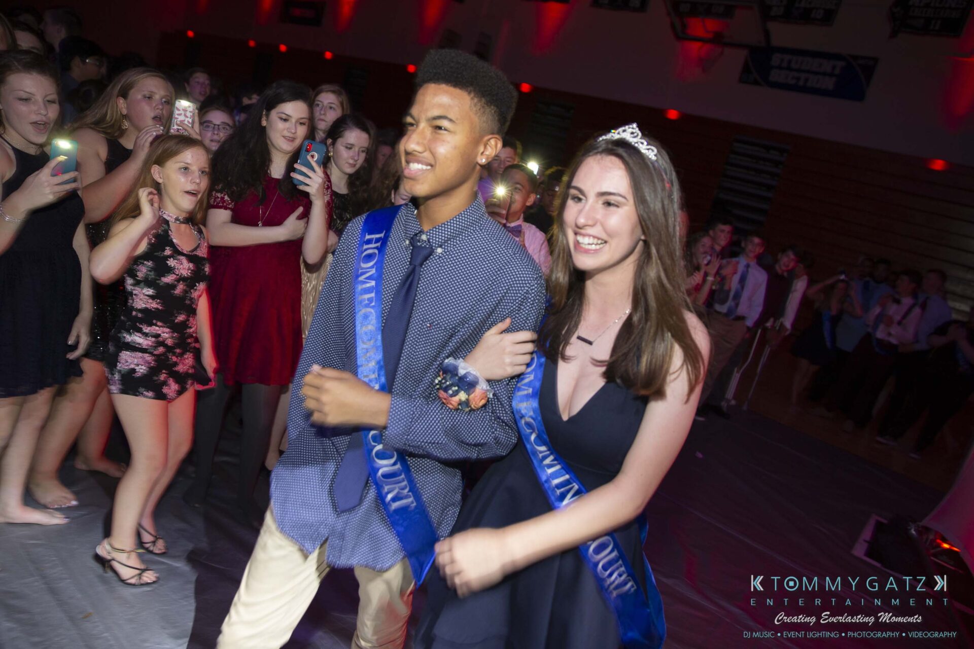 SR-Homecoming-102718-Carl-Low-Res-42 - TOMMY GATZ ENTERTAINMENT Two homecoming court members, wearing sashes, walk together at a school dance surrounded by students.