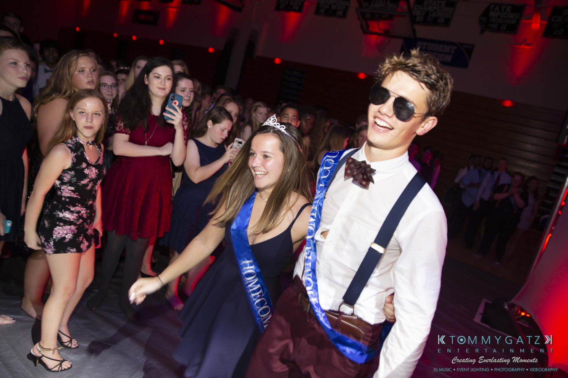 SR-Homecoming-102718-Carl-Low-Res-43 - TOMMY GATZ ENTERTAINMENT Two people wearing "Homecoming" sashes dance at a crowded event. One has a tiara and the other wears sunglasses. People in the background appear to be watching and taking photos.