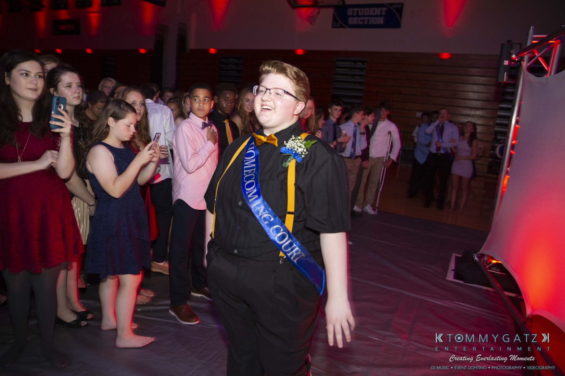 SR-Homecoming-102718-Carl-Low-Res-44 - TOMMY GATZ ENTERTAINMENT A person wearing a "Homecoming Court" sash walks confidently at a school event while others look on in a decorated hall.