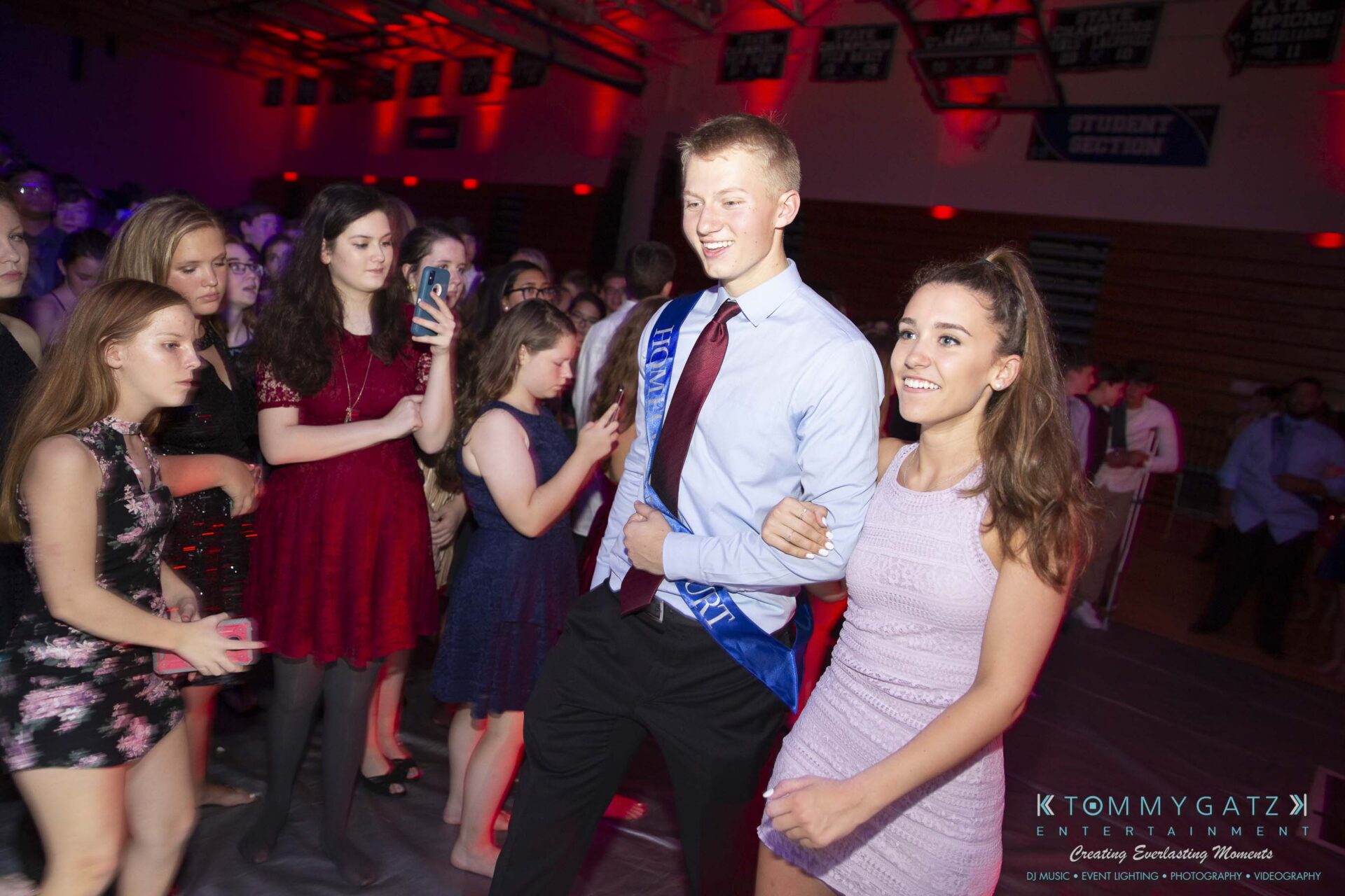 SR-Homecoming-102718-Carl-Low-Res-45 - TOMMY GATZ ENTERTAINMENT A young man and woman walk arm-in-arm on a gymnasium dance floor, surrounded by people, with a "Mister" sash worn by the man.
