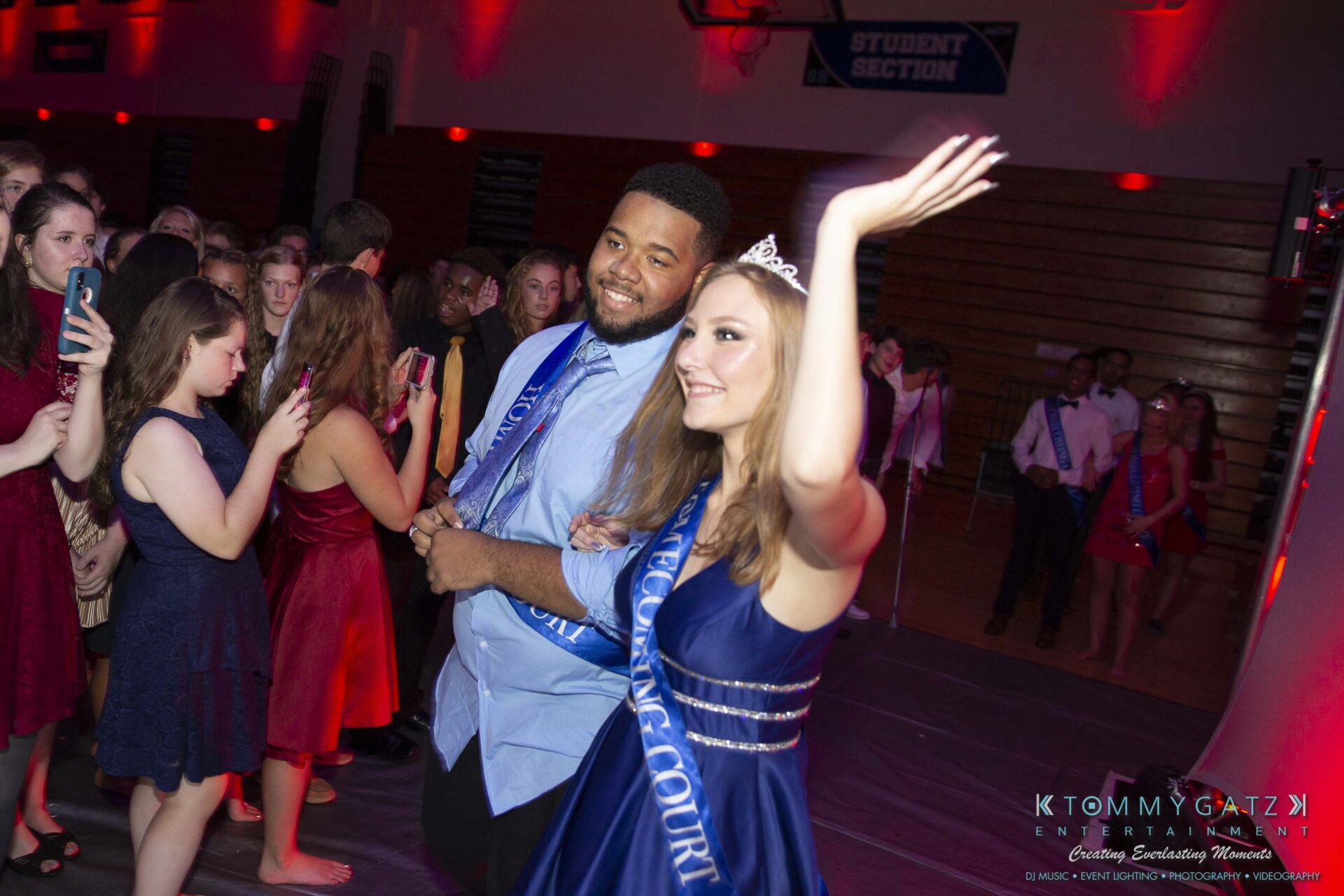 SR-Homecoming-102718-Carl-Low-Res-46 - TOMMY GATZ ENTERTAINMENT Two people wearing blue sashes stand in a gymnasium. The woman waves while wearing a tiara. A crowd of people is in the background.