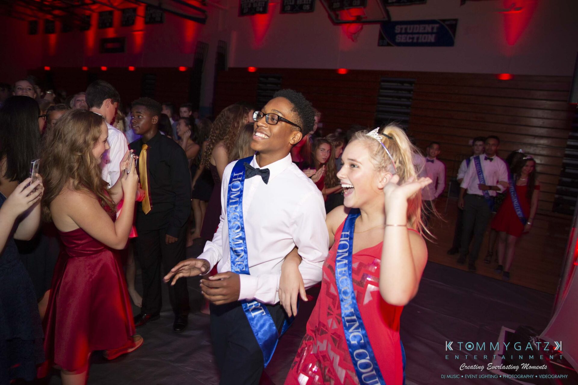 SR-Homecoming-102718-Carl-Low-Res-47 - TOMMY GATZ ENTERTAINMENT Two people wearing blue sashes smile and walk together in a gym decorated for a dance. Other attendees are visible in the background.