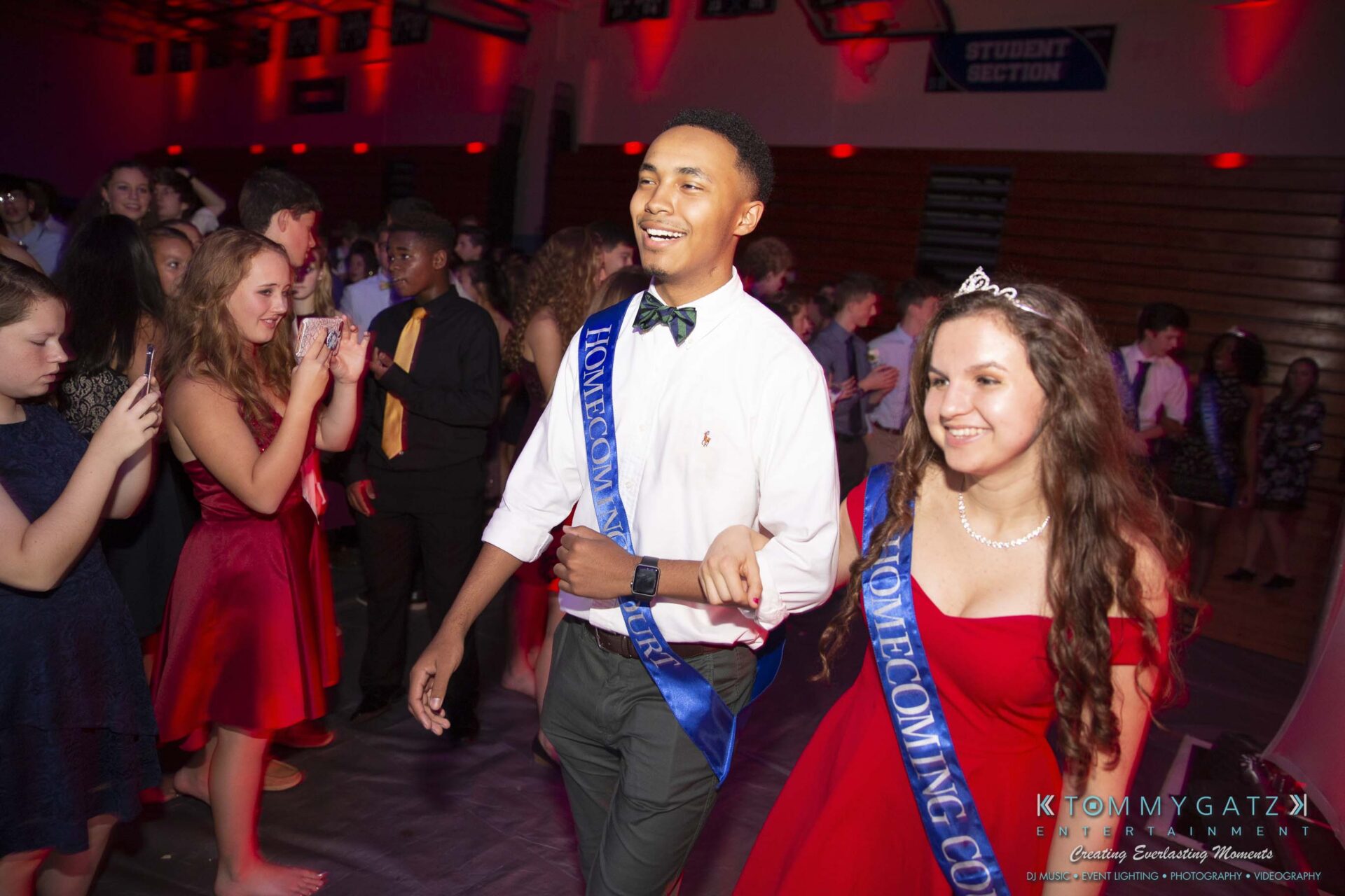 SR-Homecoming-102718-Carl-Low-Res-48 - TOMMY GATZ ENTERTAINMENT A smiling young couple wearing blue "Homecoming Court" sashes walk hand-in-hand at a school dance, with people clapping in the background.